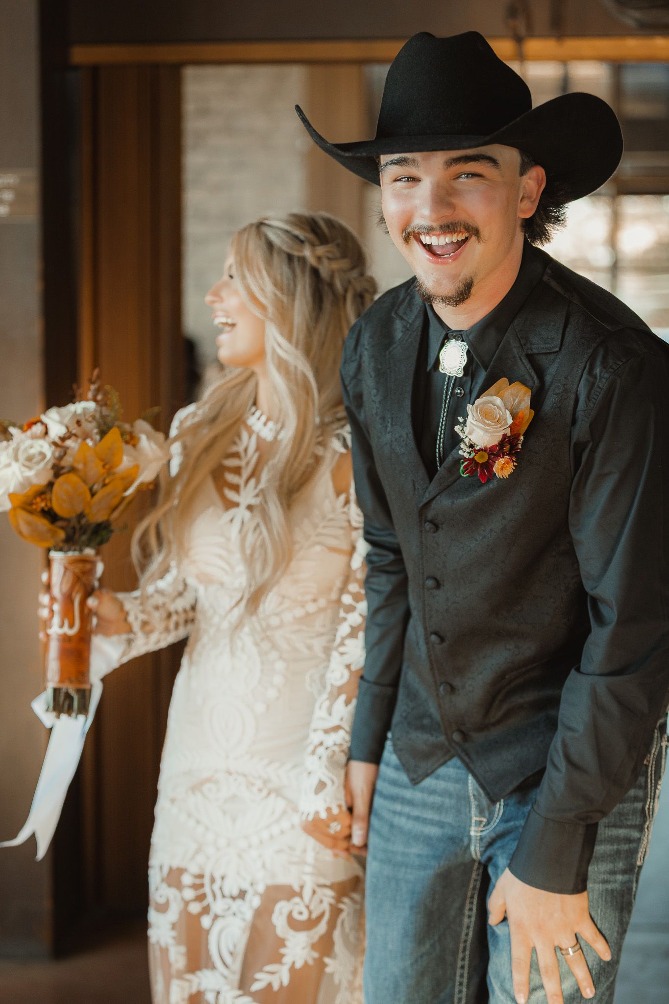 Western themed wedding couple with groom in black cowboy hat and vest standing indoors.