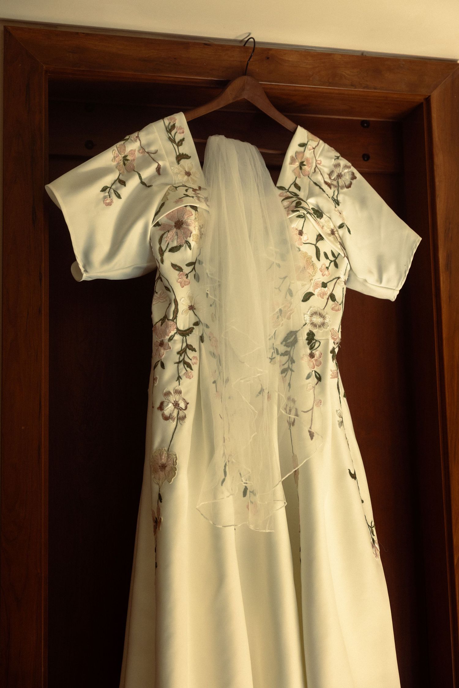 White floral embroidered wedding dress with short sleeves and veil hanging on wooden hanger in closet.