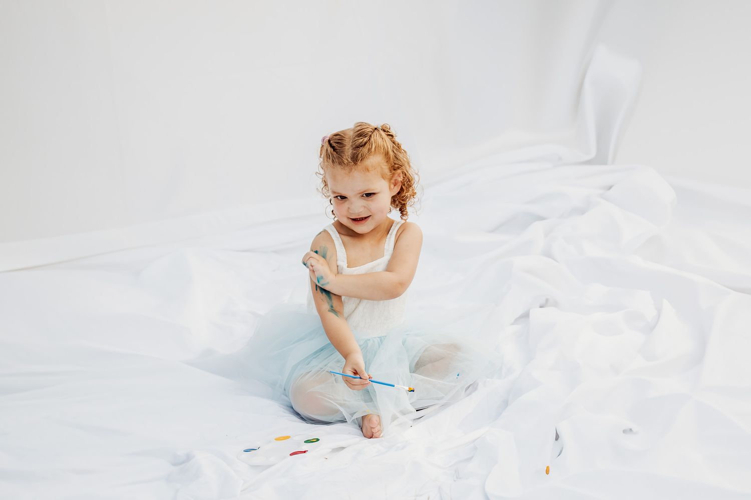 Baby in white onesie crawls and plays on soft white blanket in natural light setting.