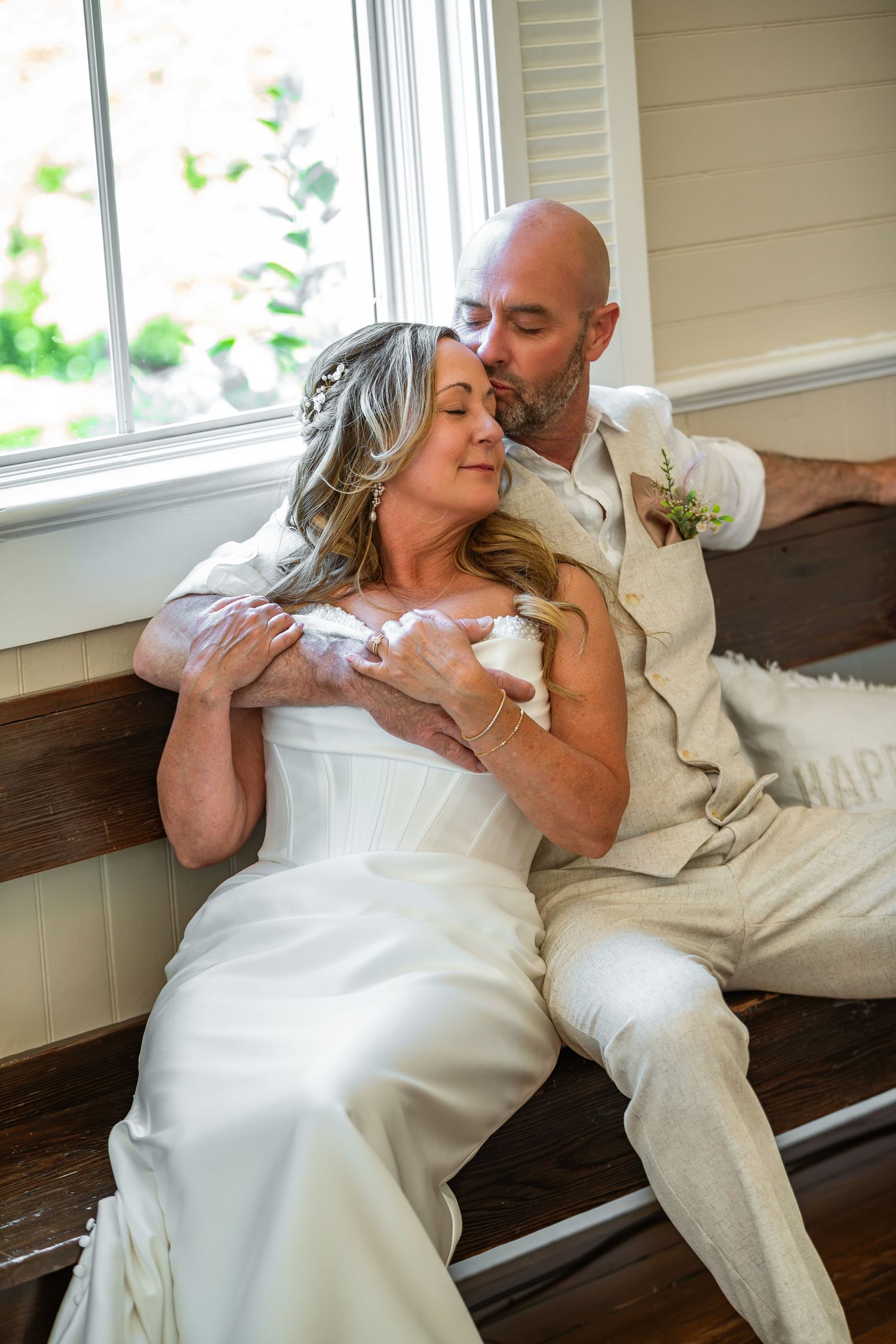 A bride and groom share a tender embrace on a wooden bench, with the groom kissing her cheek on their wedding day.