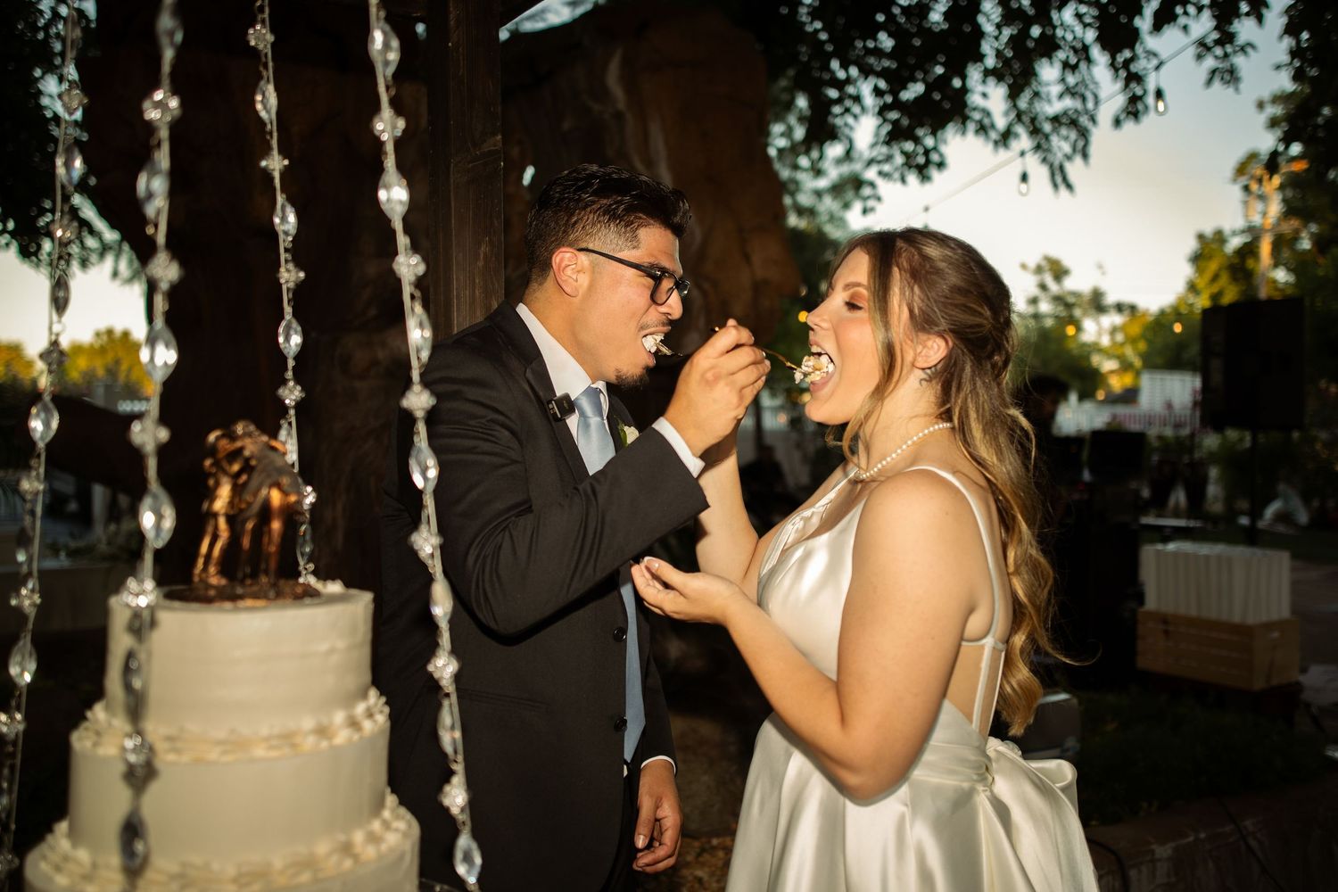 Bride and groom sharing wedding cake at outdoor reception under crystal garland decorations at dusk.