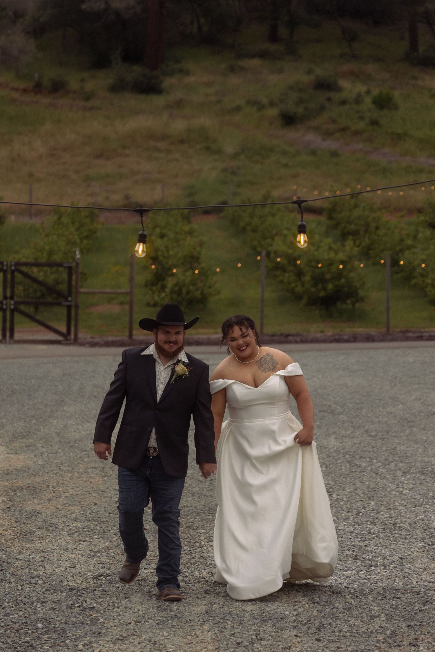 Bride in white off-shoulder gown and groom in black suit with cowboy hat walk together at outdoor wedding venue.
