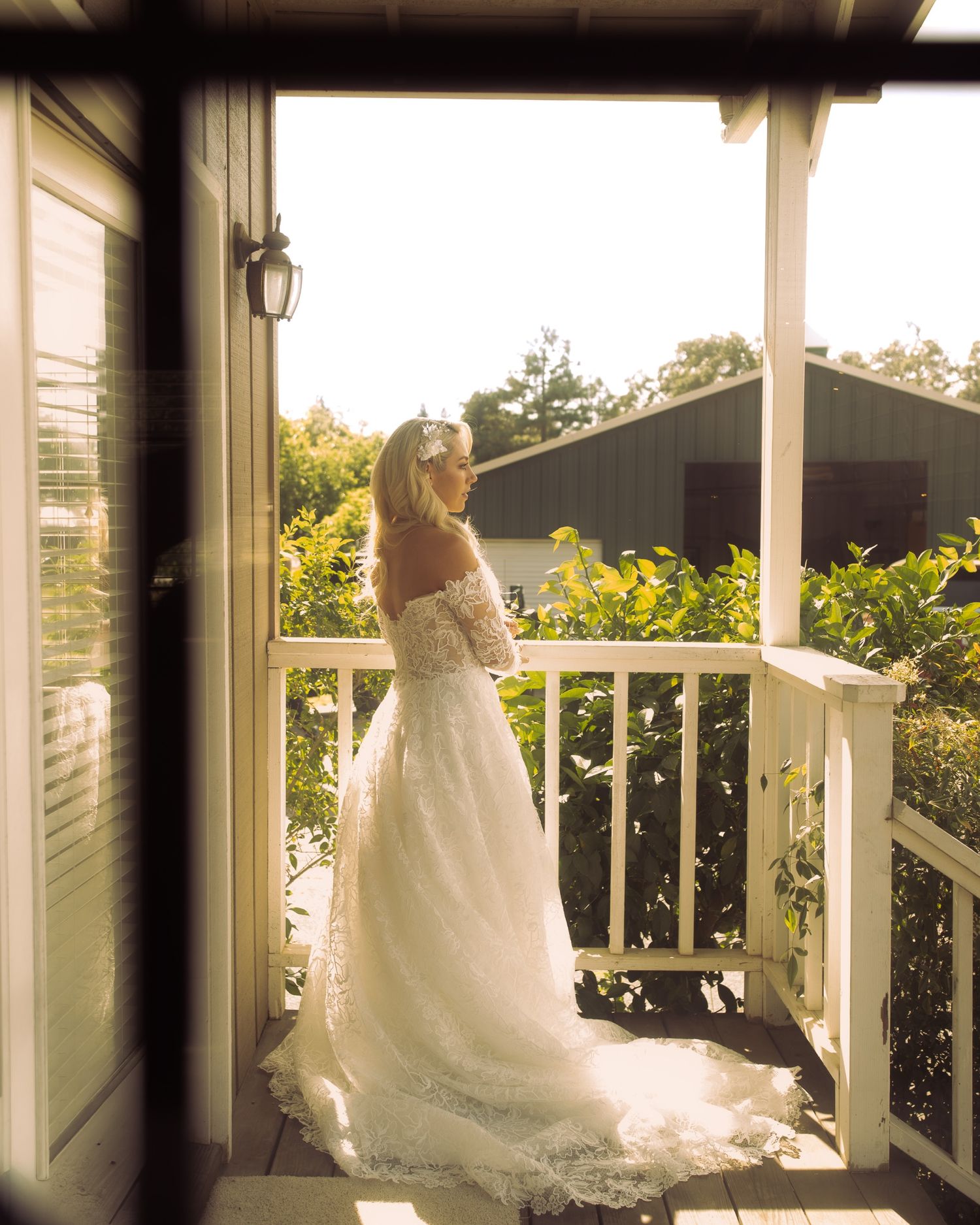 Bride in white lace off-shoulder gown stands on sunlit porch balcony surrounded by lush greenery on wedding day.