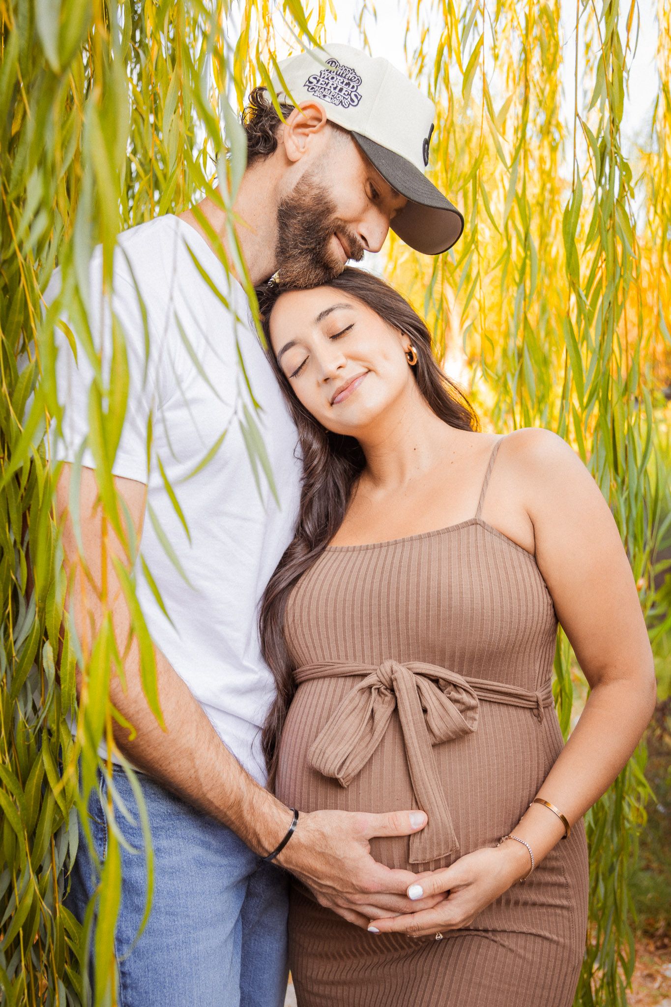 Maternity photoshoot under a willow tree with a woman in brown dress showing her baby bump in natural lighting.