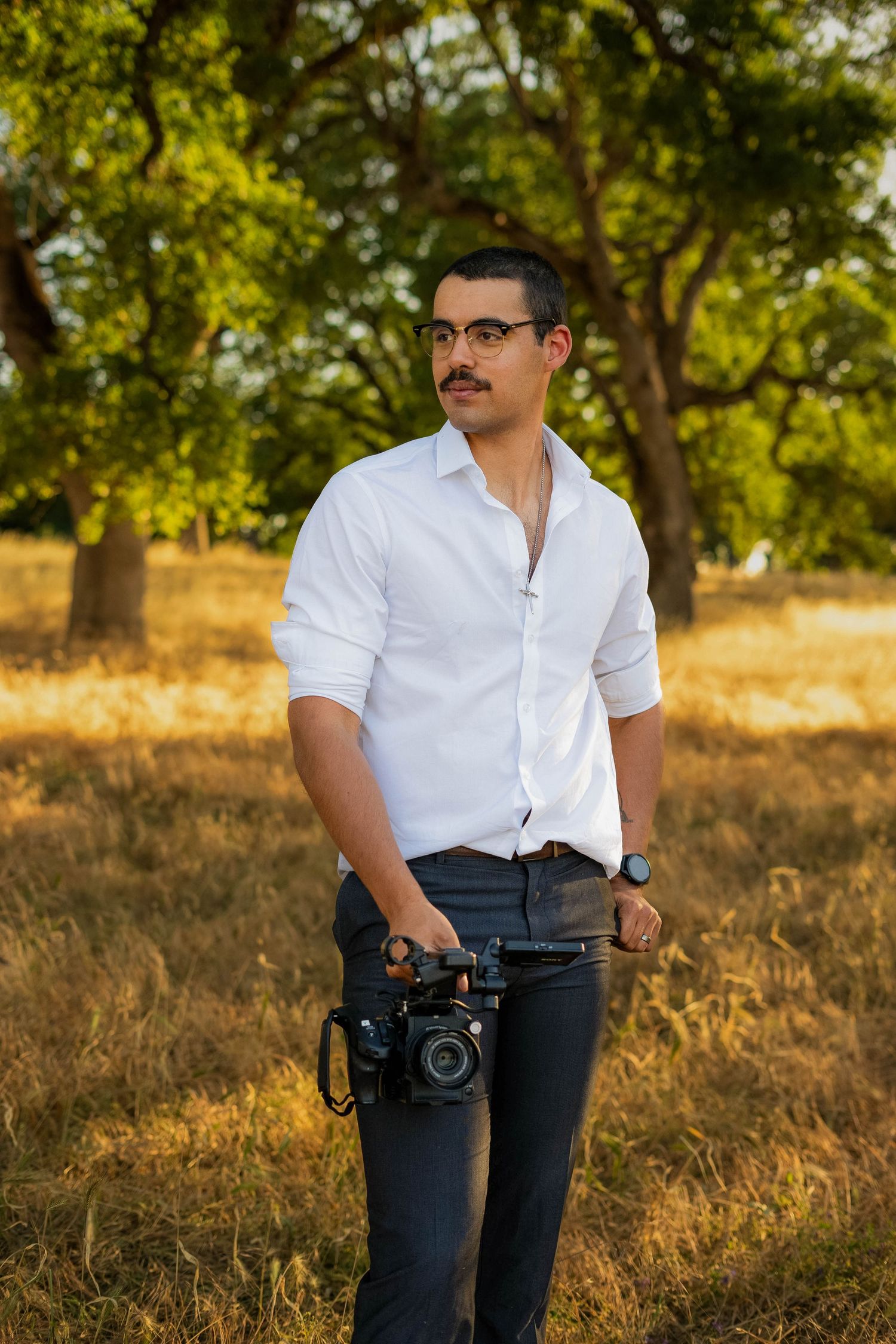 Photographer in white shirt and dark pants holding camera outdoors in golden sunlit field with trees.