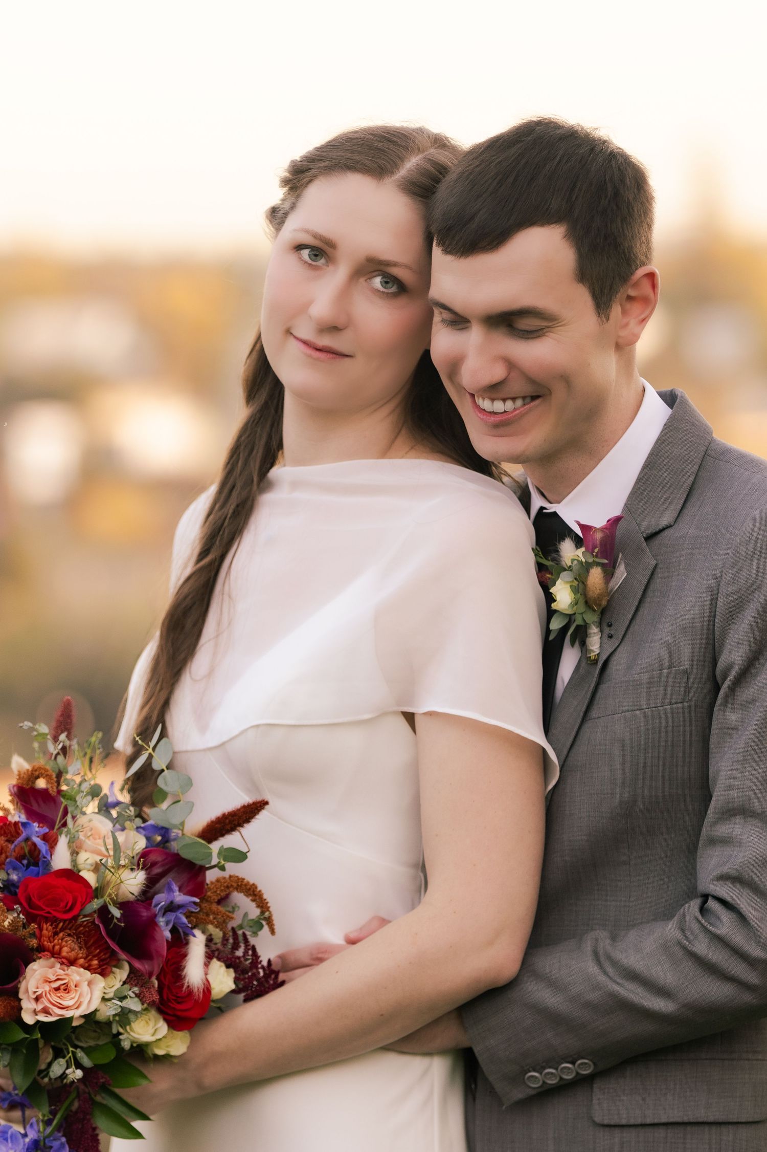 Couple in wedding attire share intimate moment at sunset with burgundy and blush floral bouquet.