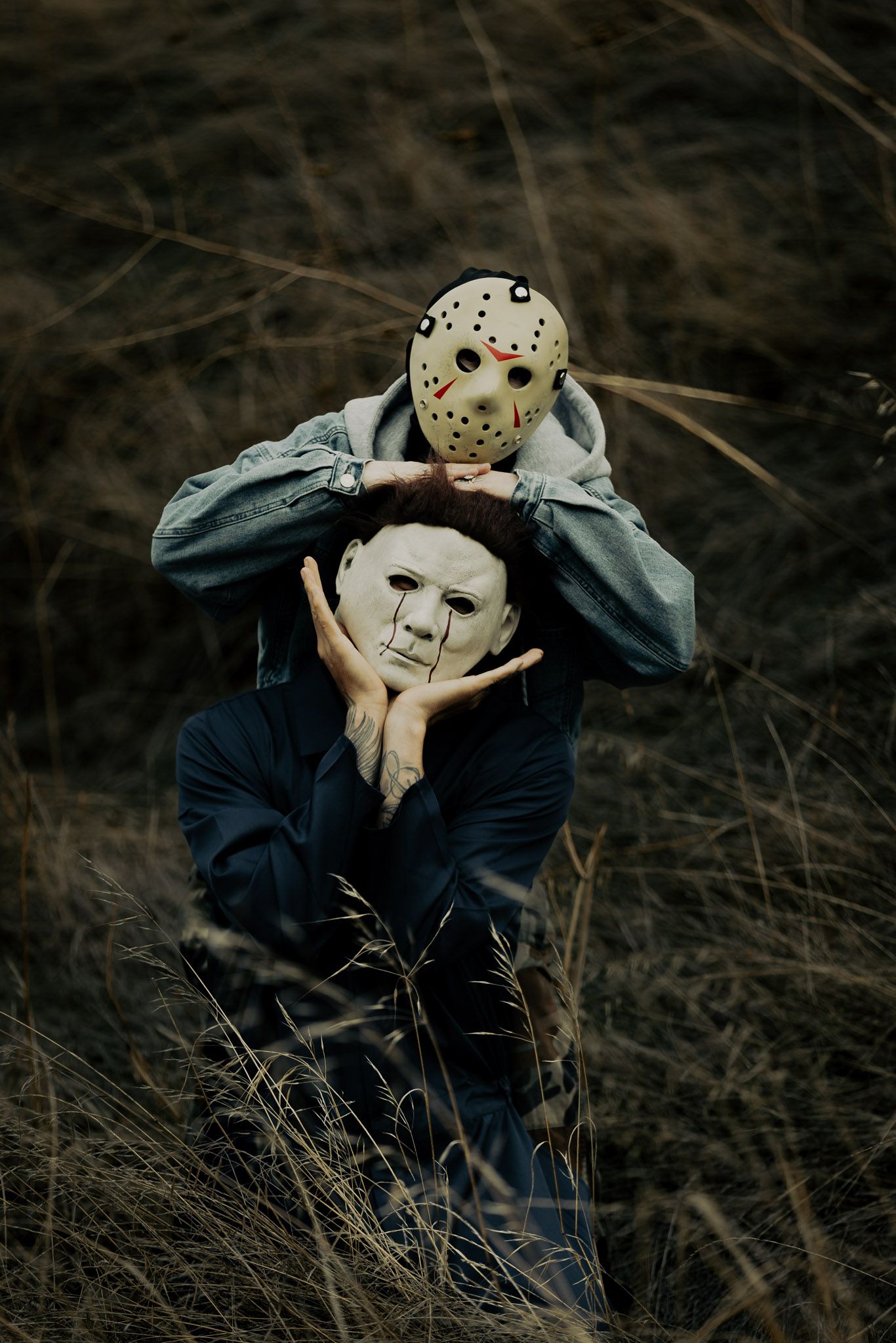 Two figures in dark clothing embrace while one wears a white hockey mask in a moody outdoor setting.