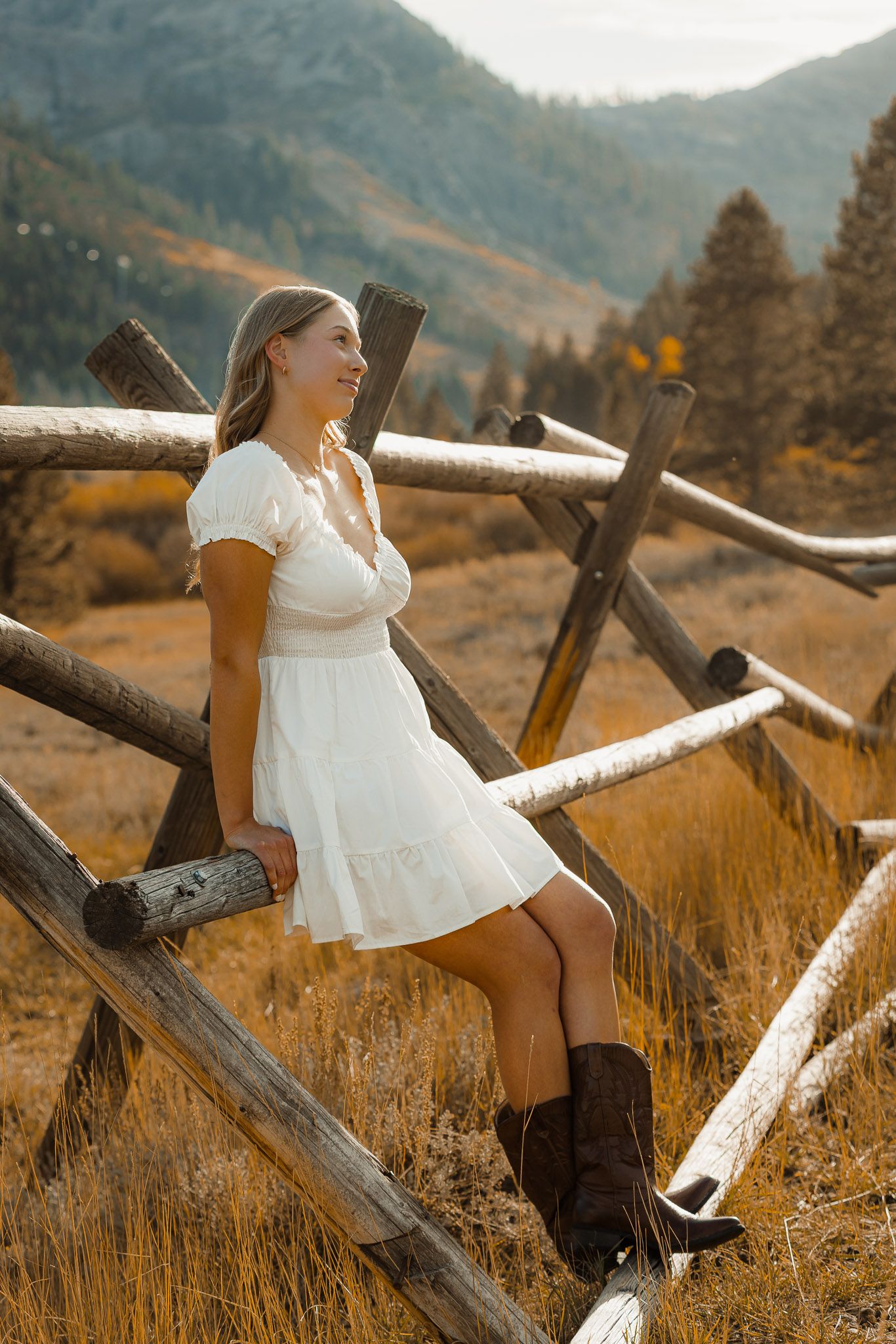 A person in a white dress sits on rustic wooden fence posts against an autumn mountain landscape.