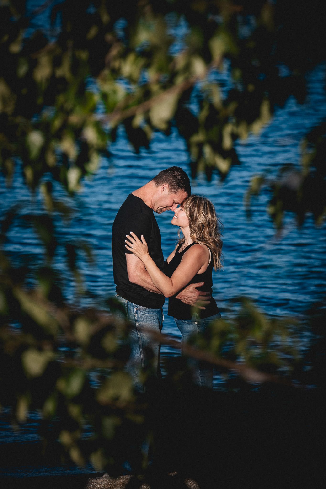 Romantic couple embraces near a serene lake with blue water and tree branches framing the scene.