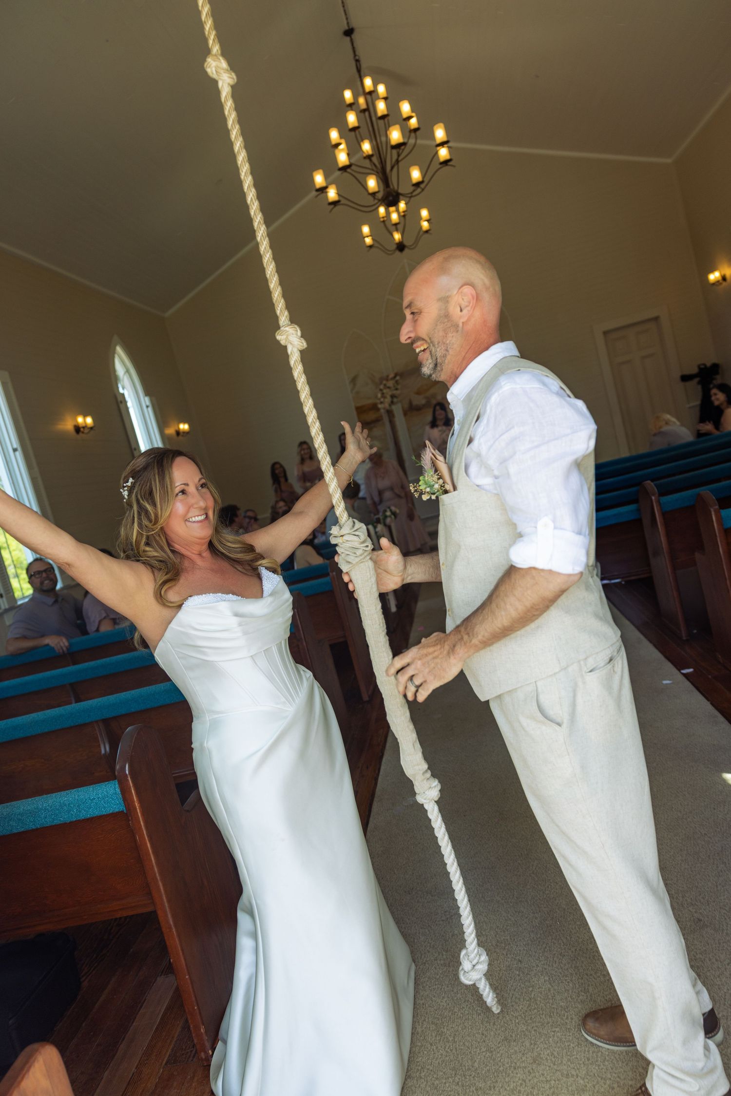 Bride and groom joyfully pulling a rope together inside a charming chapel with wooden pews on their wedding day.