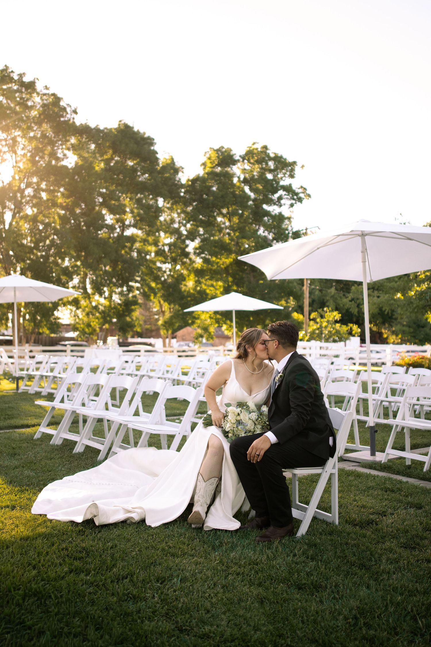 Bride and groom share a kiss while seated on white chairs at an outdoor wedding ceremony surrounded by lush green trees.