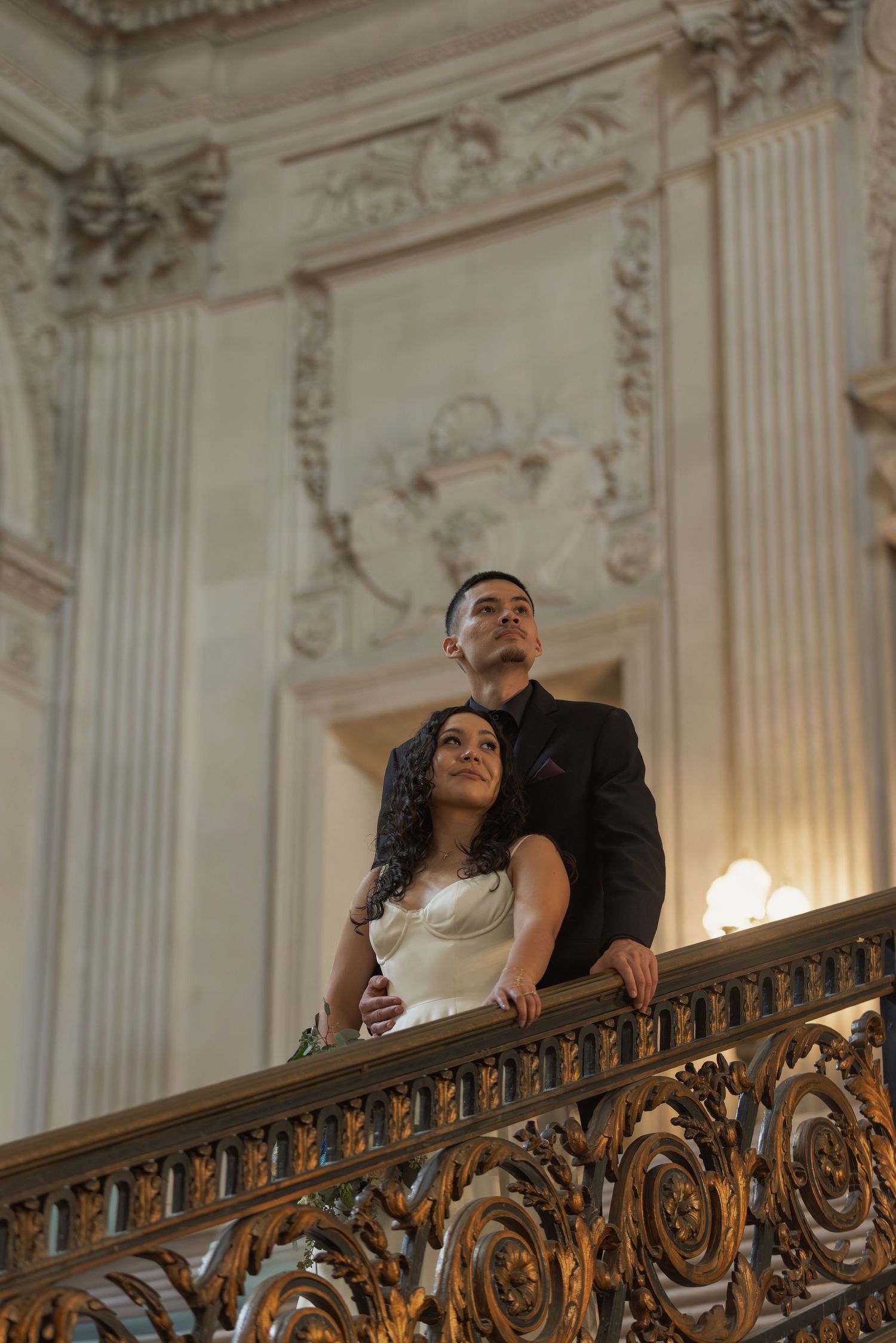 Elegant couple in wedding attire poses on ornate golden staircase railing in grand classical architecture venue.