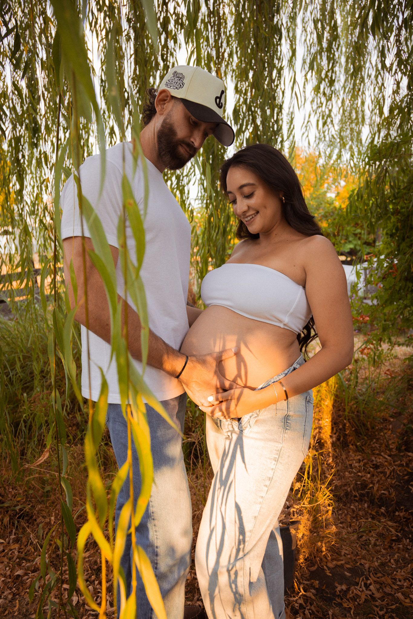 A couple shares a tender moment under a willow tree during a maternity photoshoot at sunset.