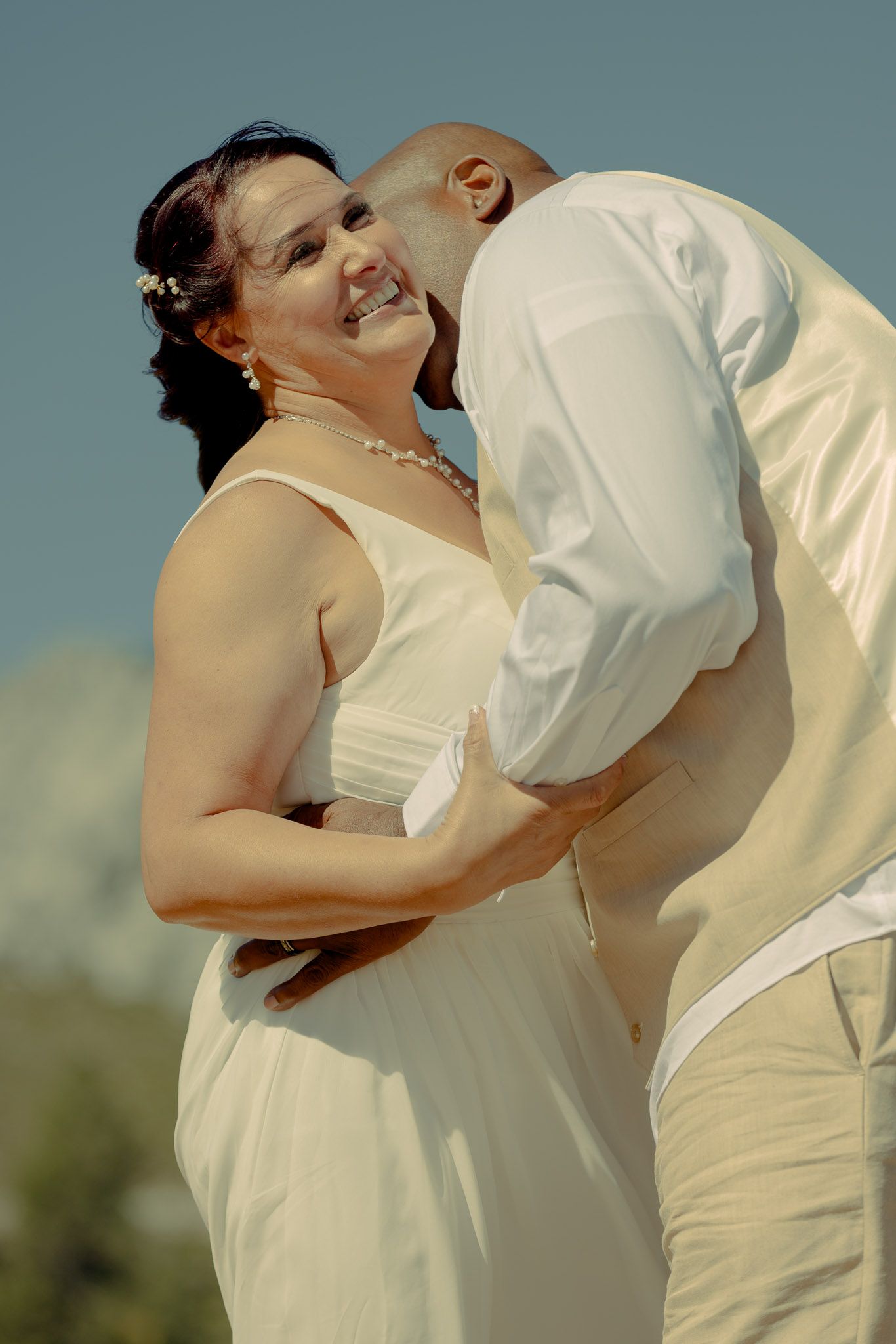 Couple in white attire sharing romantic moment against blue sky background.