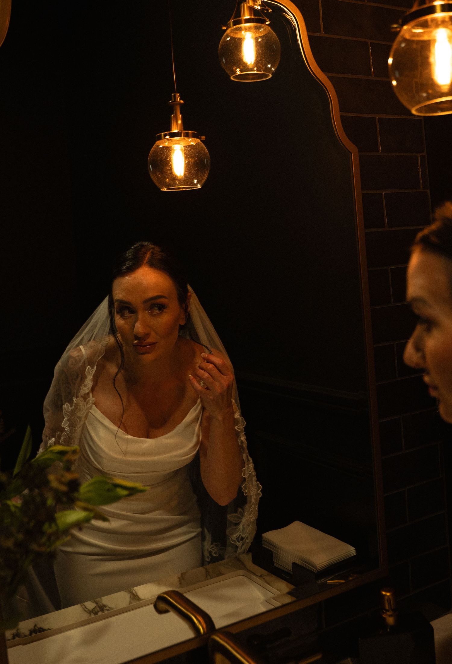 Bride in white gown and lace veil adjusts her earring before a gold-framed mirror in a moody, warmly lit bathroom.