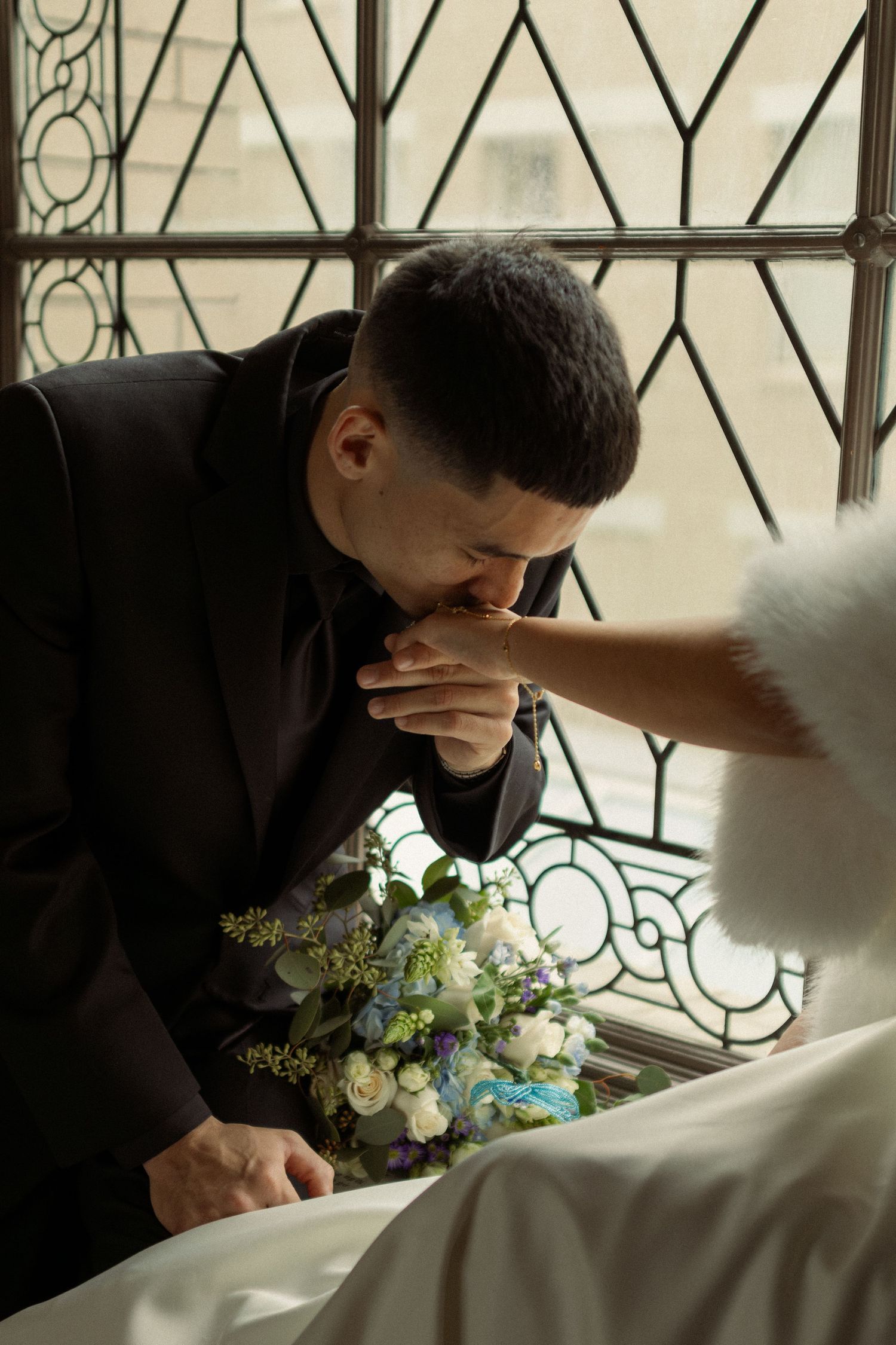 Groom in black suit kissing bride's hand during wedding ceremony, with bouquet of blue and white flowers nearby.