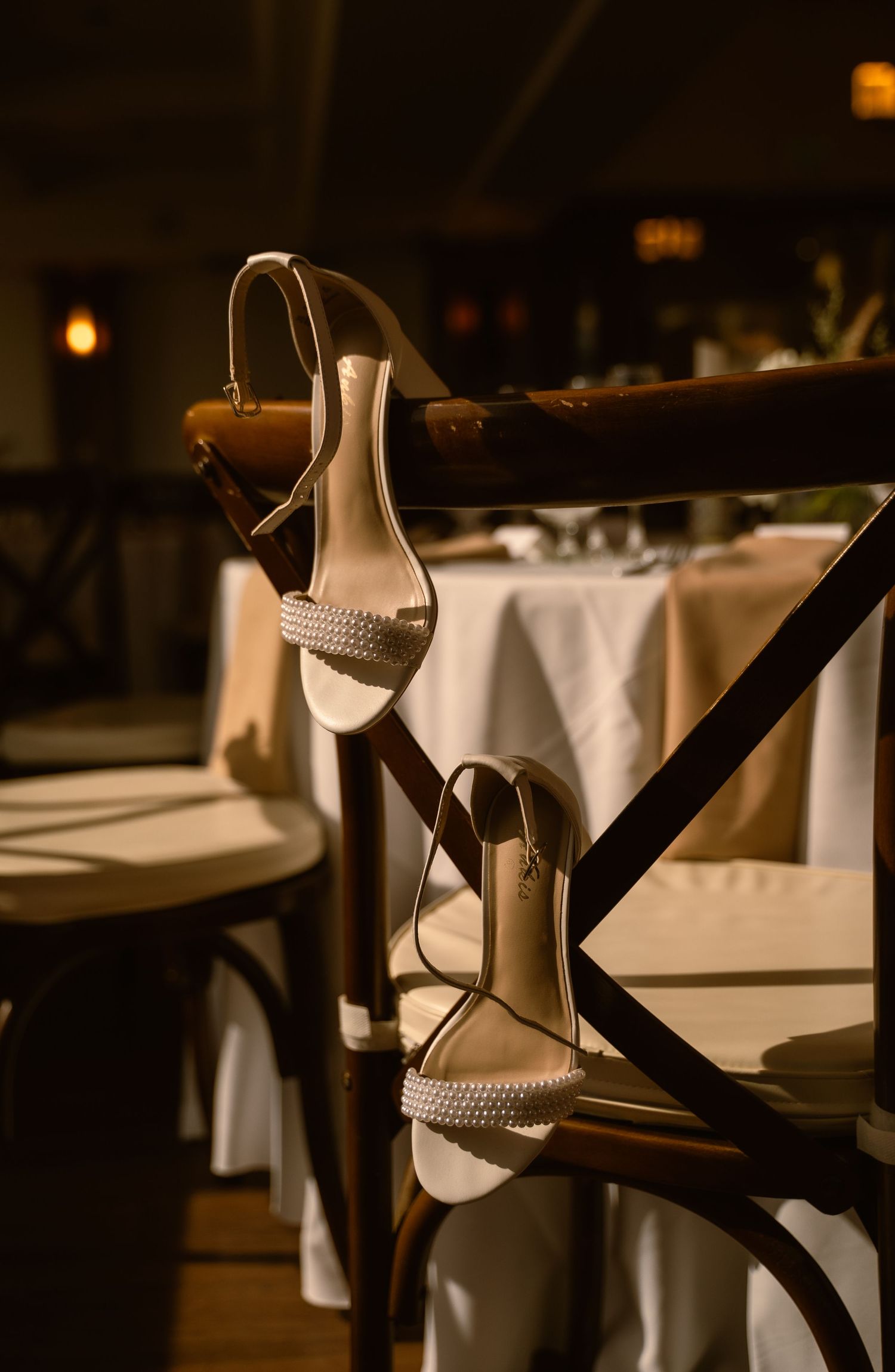 Elegant white strappy heeled sandals hanging on a wooden cross-back chair in a softly lit restaurant setting.