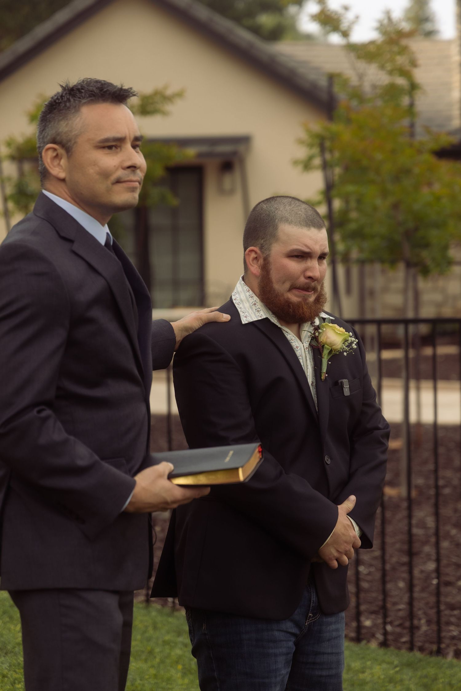 Two men in suits stand outdoors at a wedding ceremony, one holding a Bible and comforting the emotional groom.