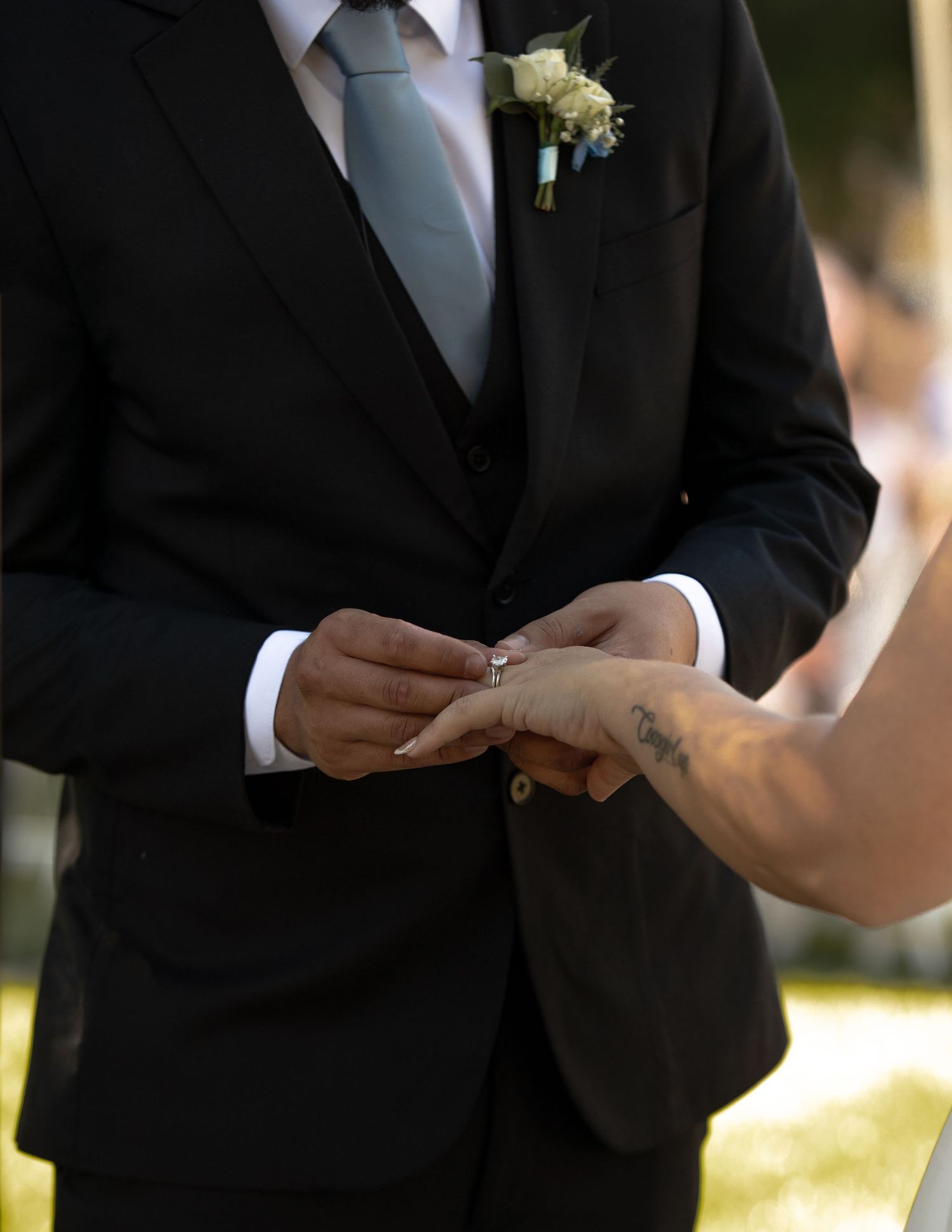 Groom in black suit placing wedding ring on bride's hand during outdoor ceremony.