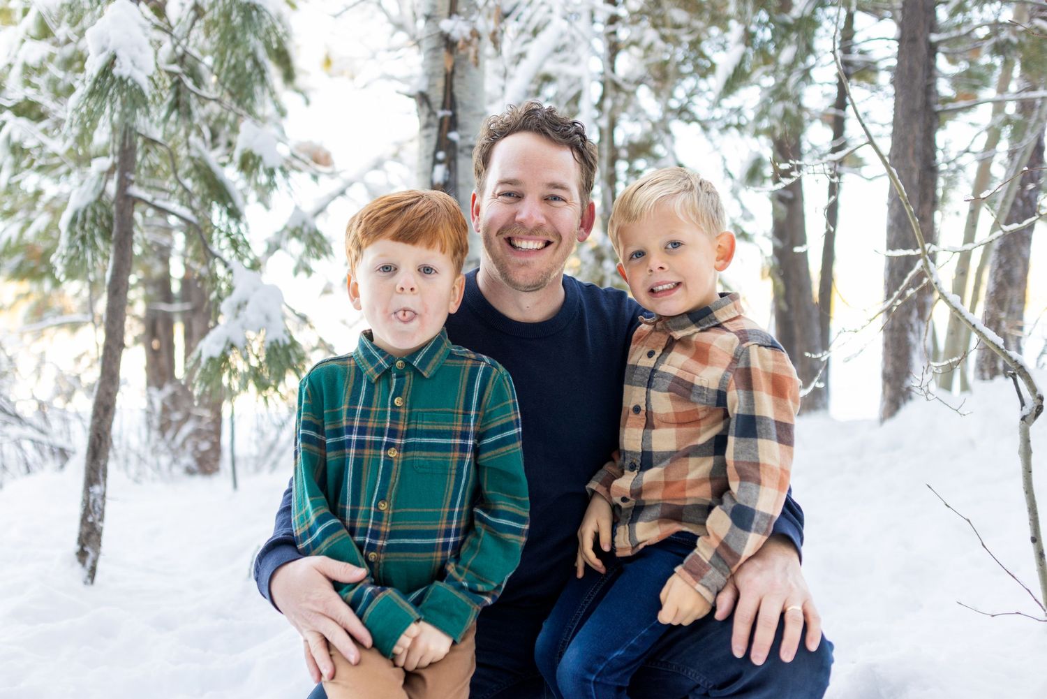 A family wearing coordinated green plaid and navy outfits poses together in a snowy winter forest setting.