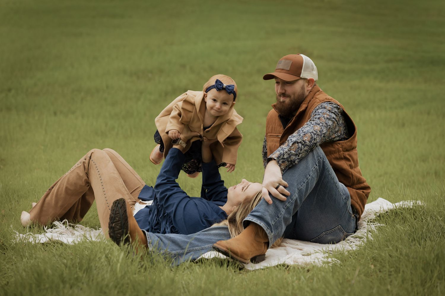 Family cuddles together while lying on blanket in green grass during outdoor portrait session.