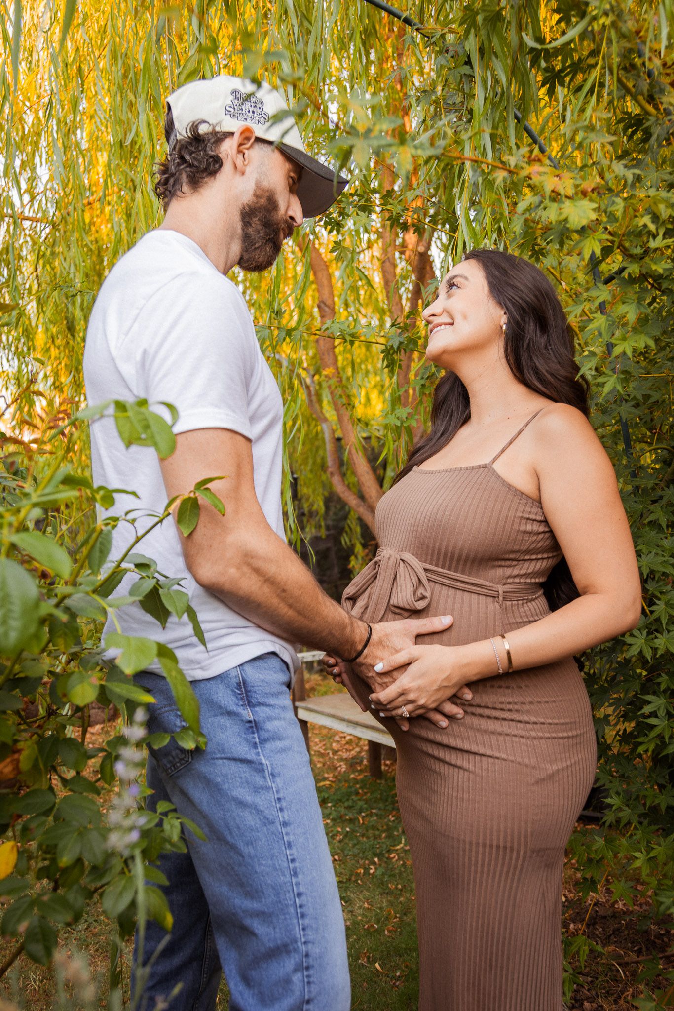 Expecting couple shares a tender moment under a willow tree in a garden setting.
