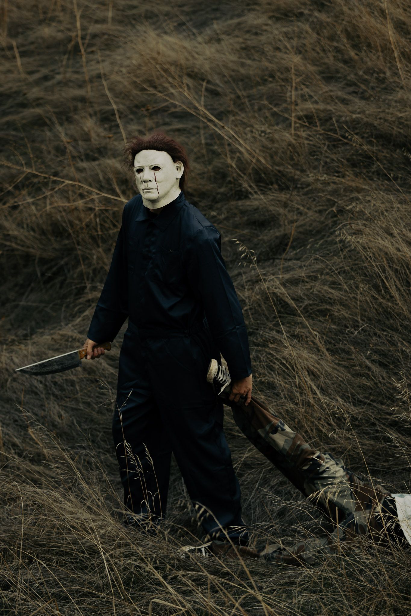 A figure in black clothing sits among dried grass while wearing a white mask.