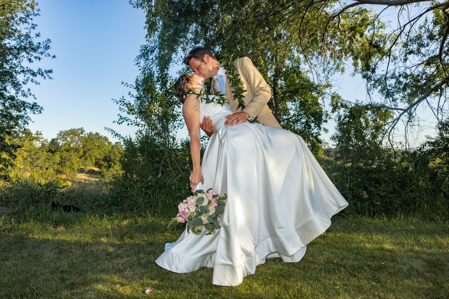 Bride and groom sharing a romantic kiss outdoors under a tree on their wedding day, bride holding a pink bouquet.