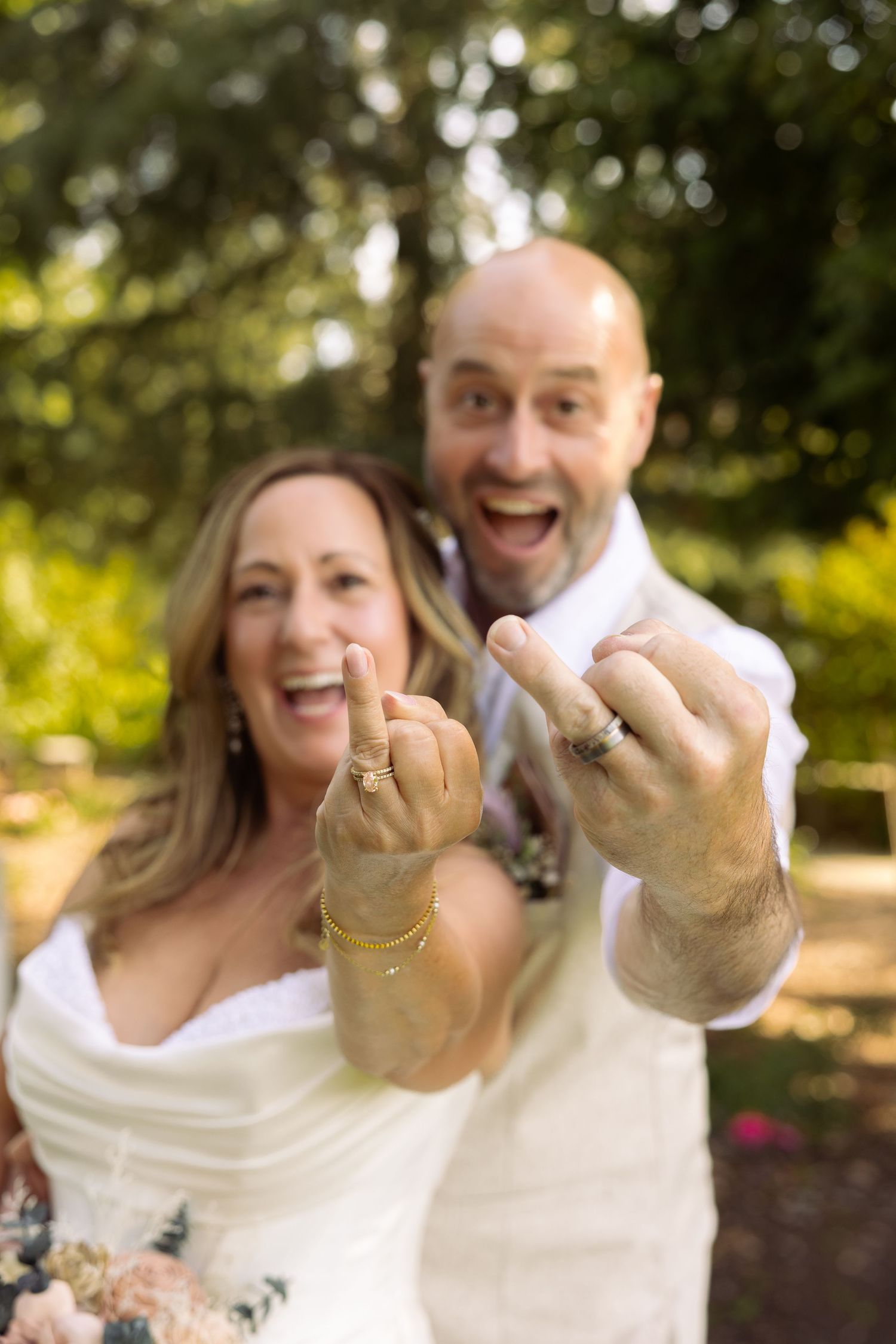 Joyful newlyweds showing off wedding rings outdoors, laughing couple in wedding attire celebrating marriage.