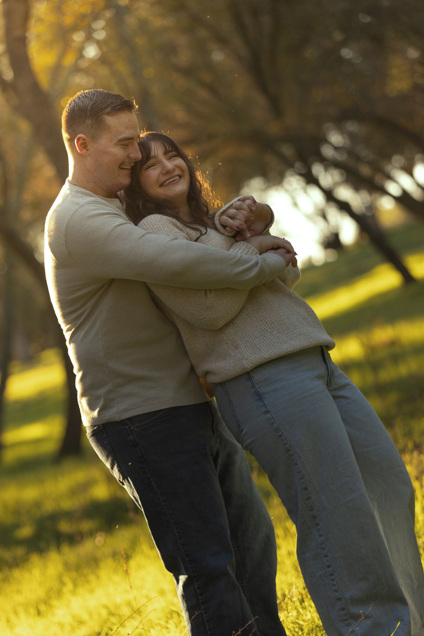 A couple embraces and laughs together in a sunlit park during golden hour with autumn trees in the background.