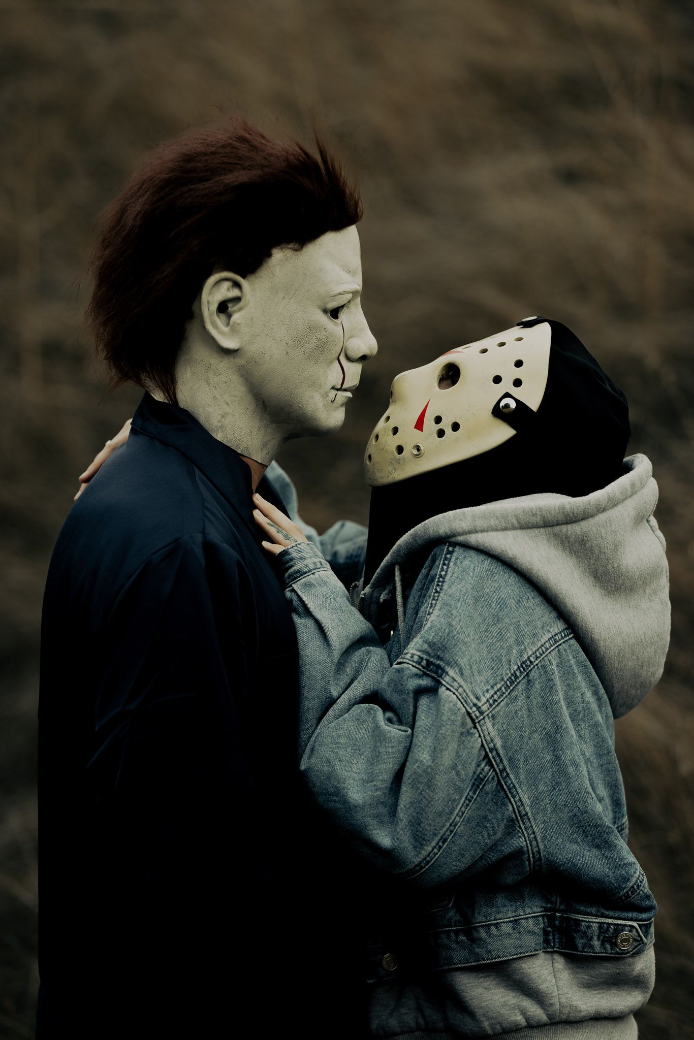 Two figures wearing denim and dark clothing embrace in a moody outdoor setting.