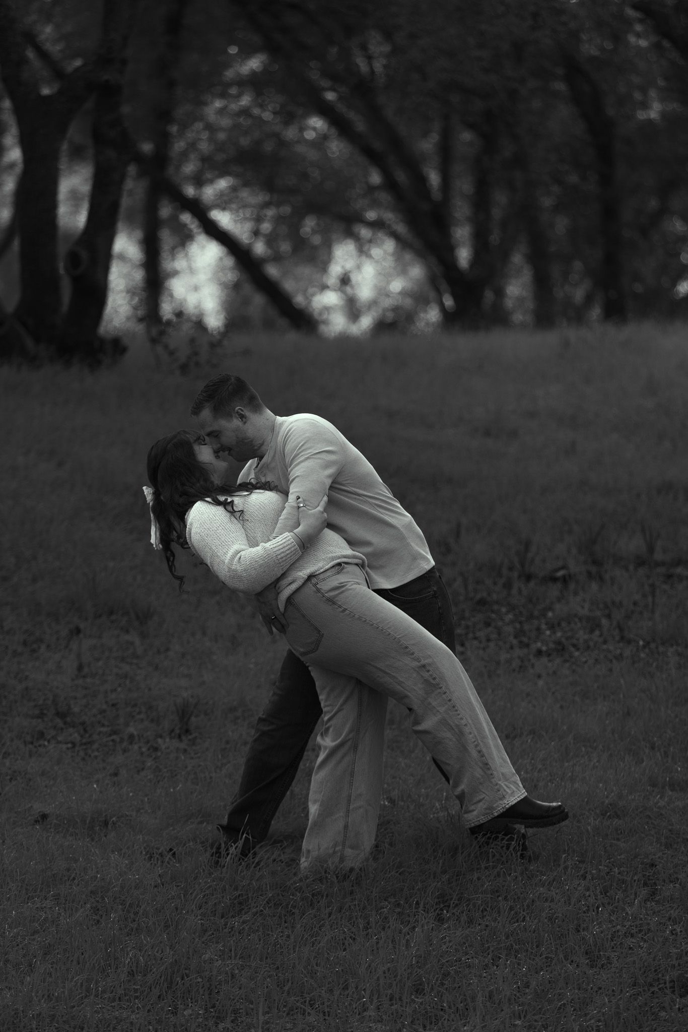A person in white clothing crouches down in a grassy field with trees in the background, creating a moody black and white scene.