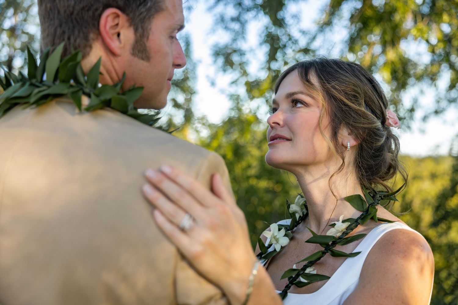 A bride in a white dress gazes lovingly at her groom wearing a Hawaiian lei during an outdoor wedding ceremony.