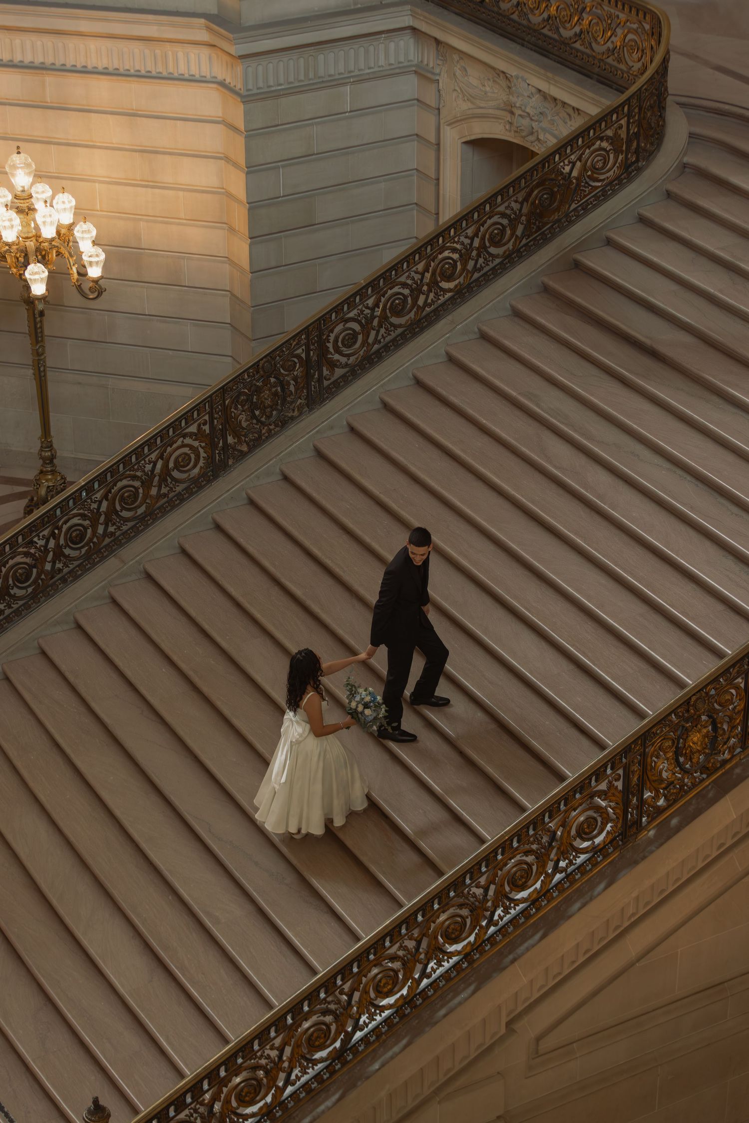 Bride and groom hold hands on ornate grand staircase with gold railings at San Francisco City Hall wedding.