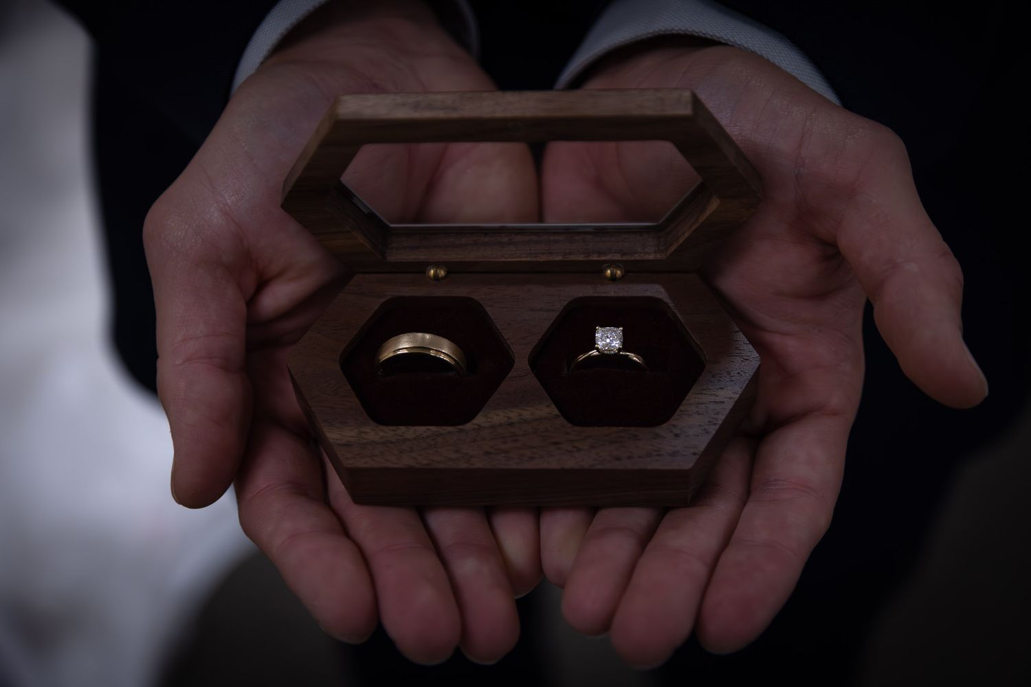 A wooden ring box containing two wedding rings held in cupped hands against a dark background.