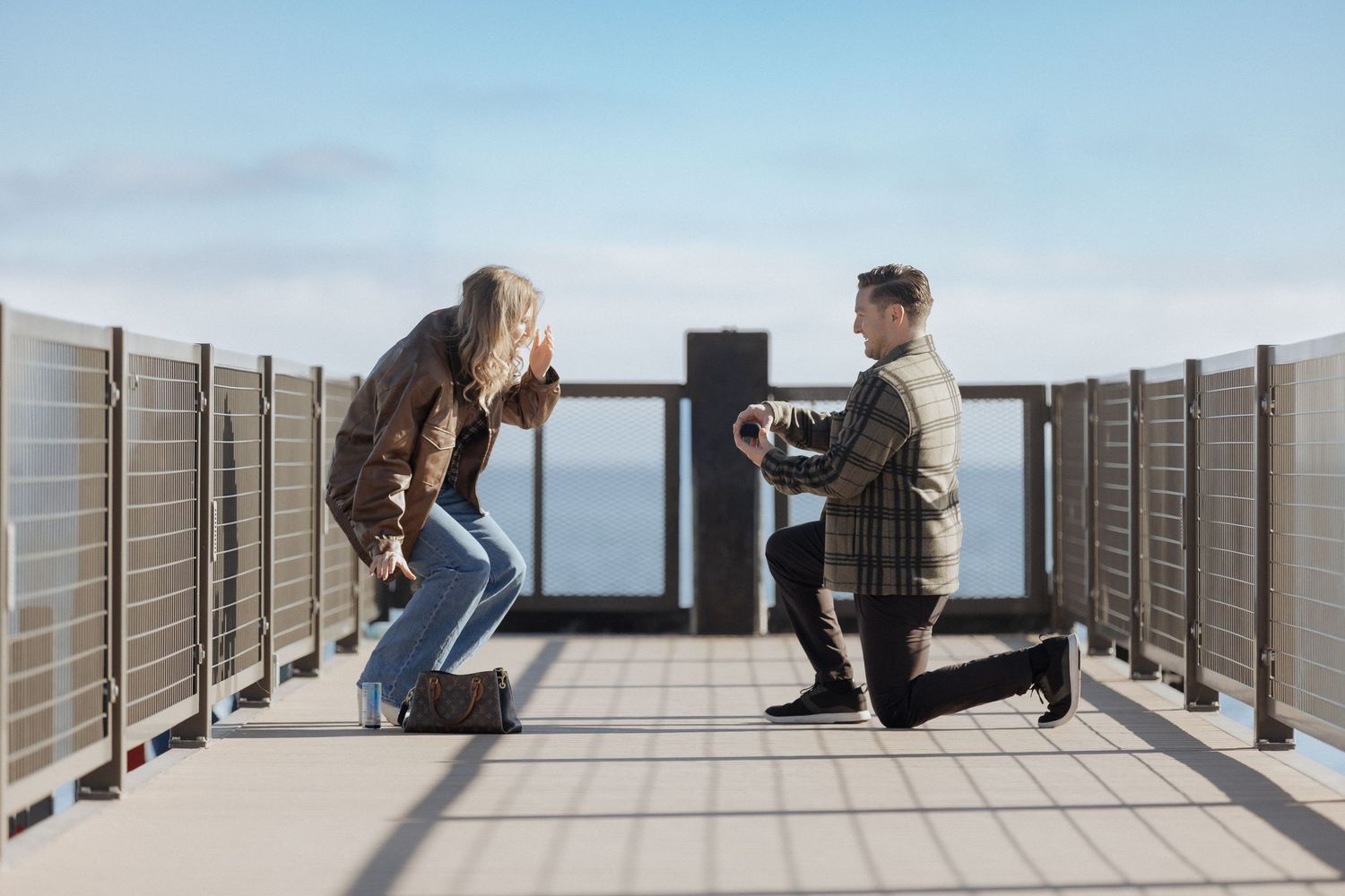 Person proposing to another on an outdoor deck overlooking a scenic view on a sunny day.