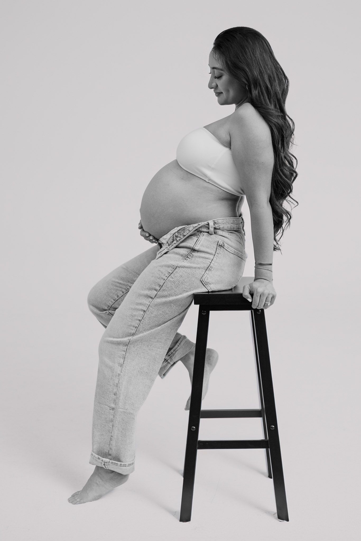 A pregnant woman in casual attire poses on a wooden stool against a gray studio backdrop.
