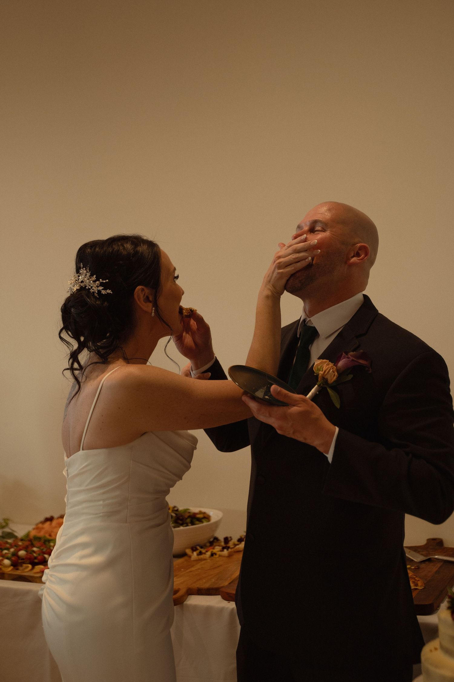 Bride and groom feed each other wedding cake at their reception near a dessert table.