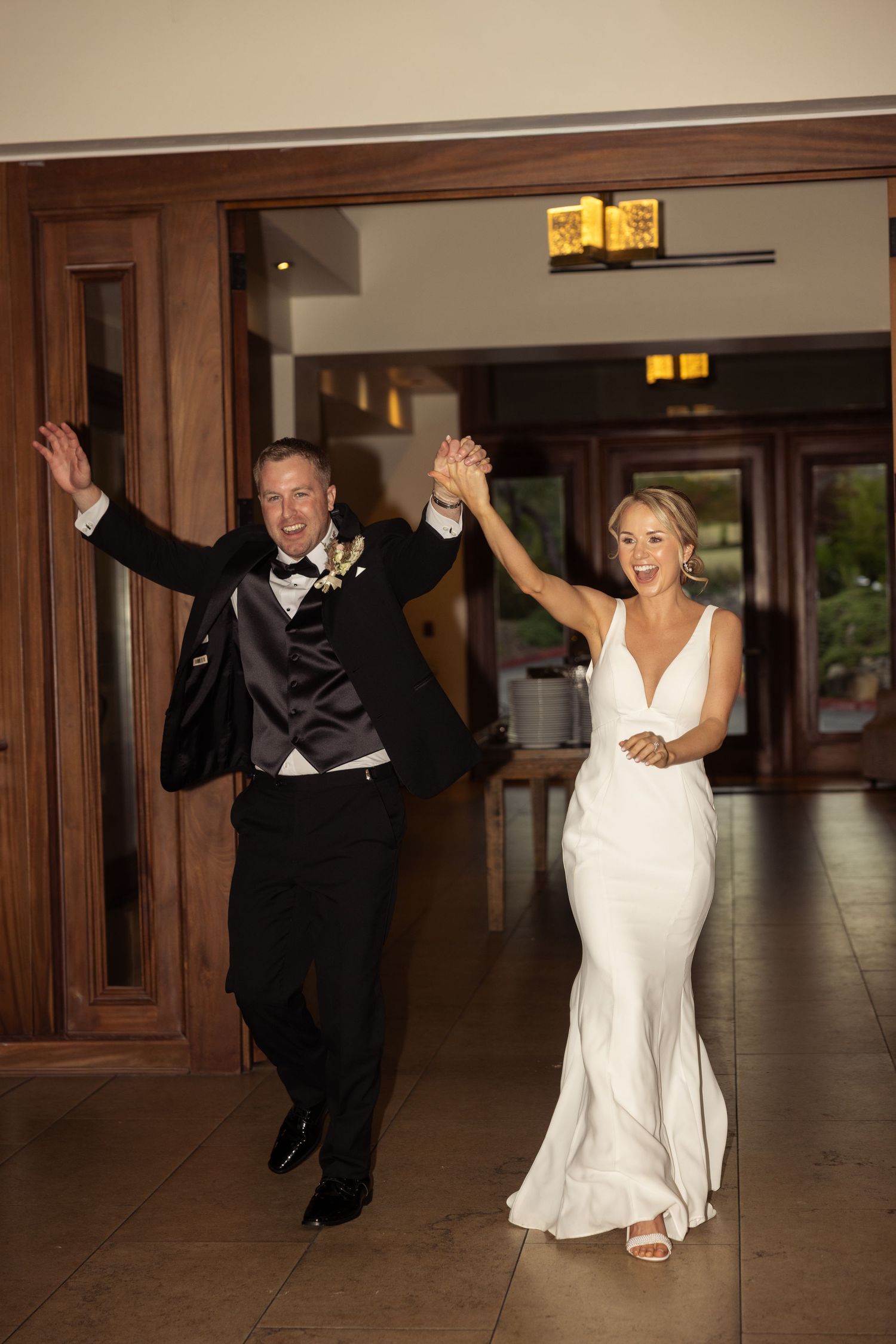 Joyful bride in white gown and groom in tuxedo holding hands, making grand entrance at wedding reception.