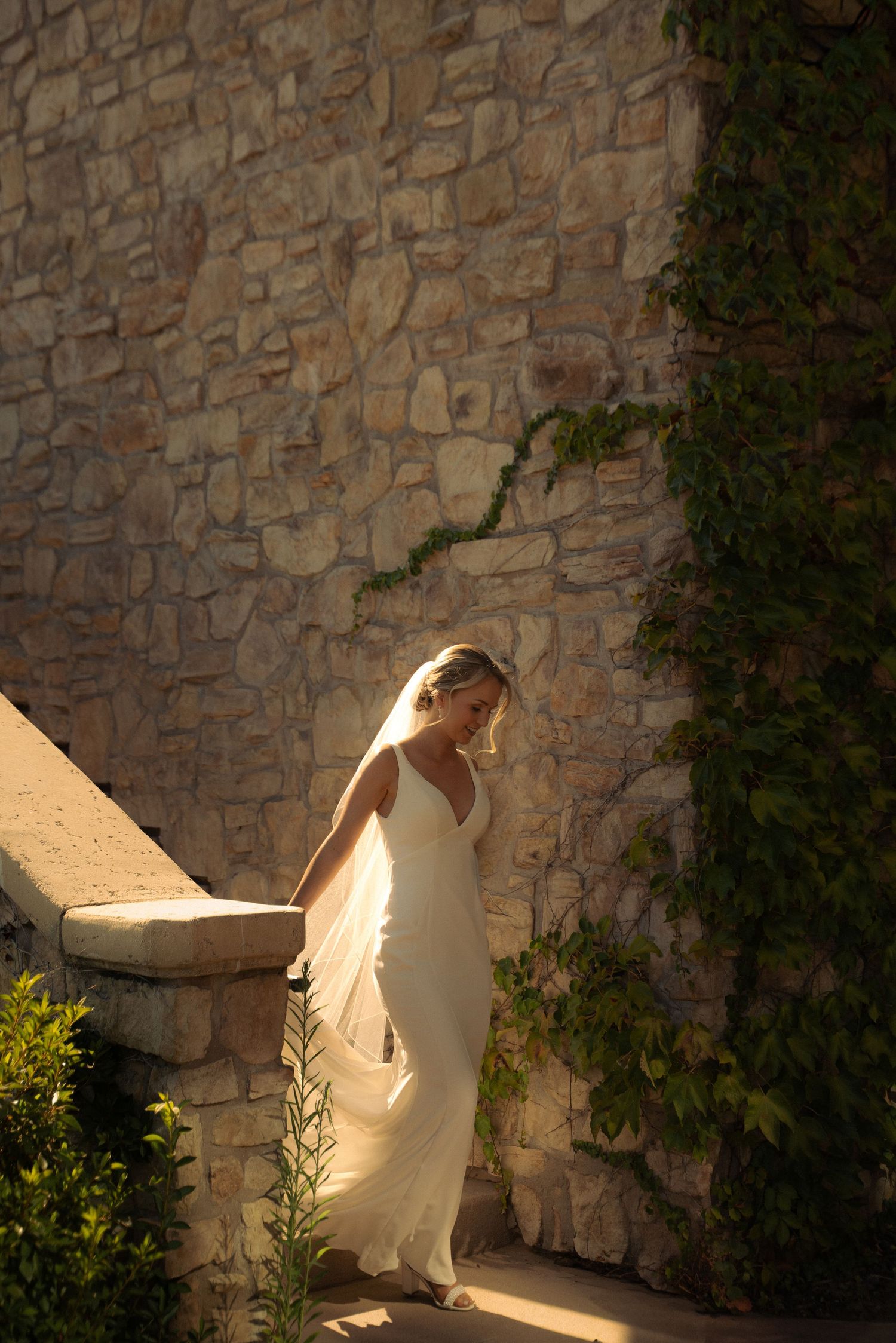 Bride in white gown and veil stands on stone steps beside ivy-covered wall in golden sunlight.