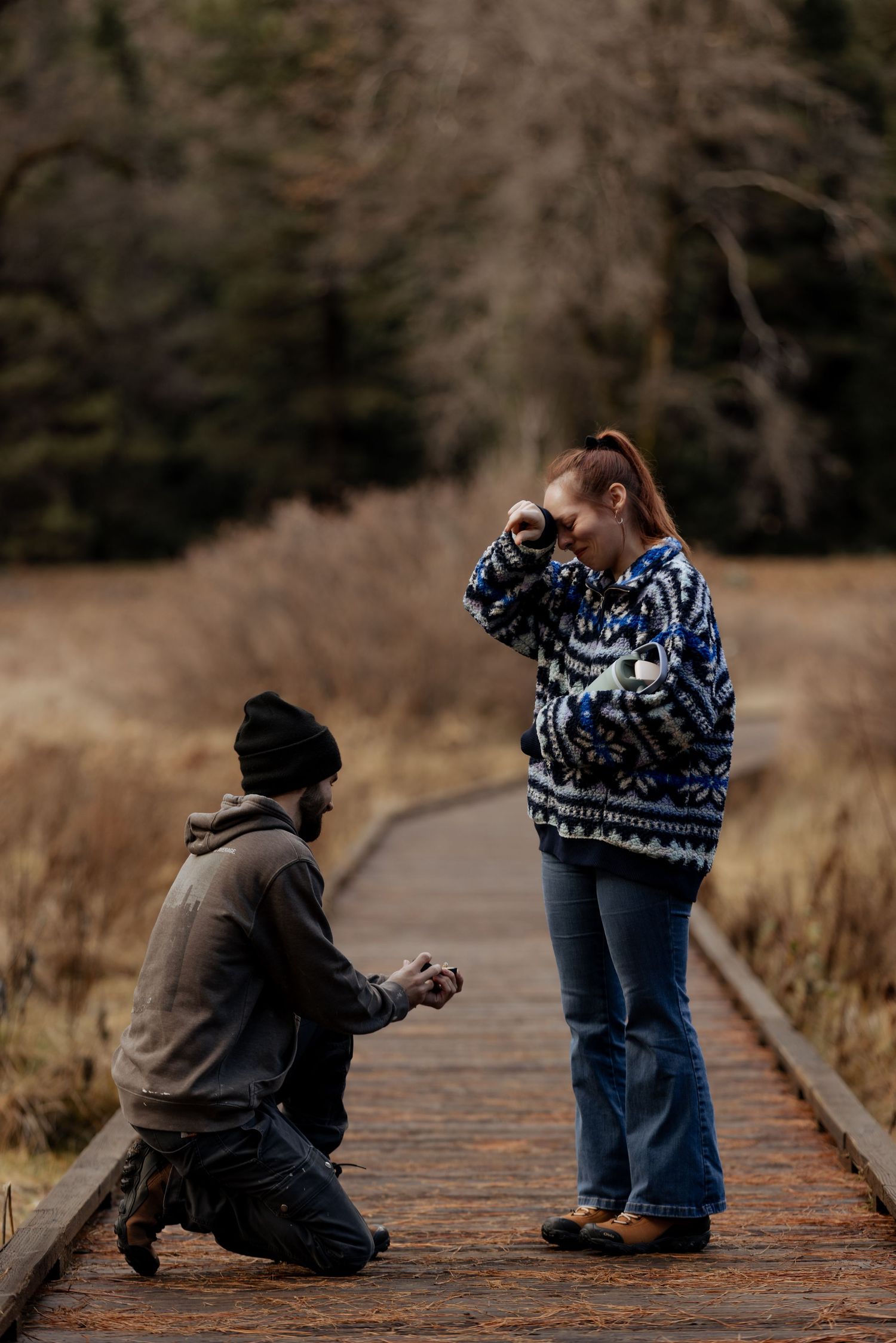 Two people sit apart on a wooden boardwalk in a natural setting during autumn.