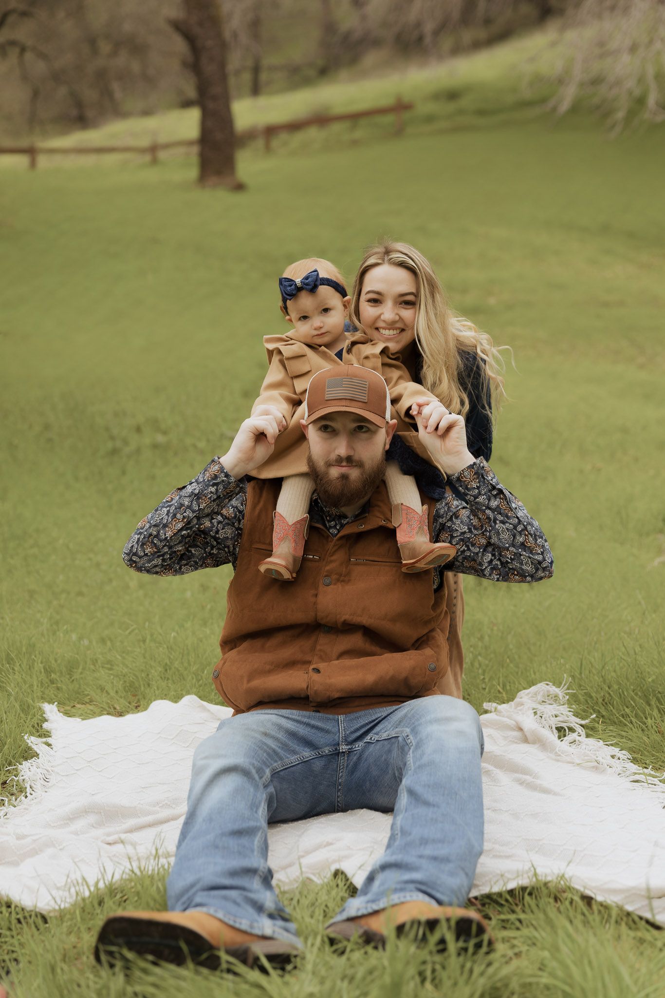 Person sitting on white blanket in grassy field wearing brown jacket and jeans while making playful expressions.