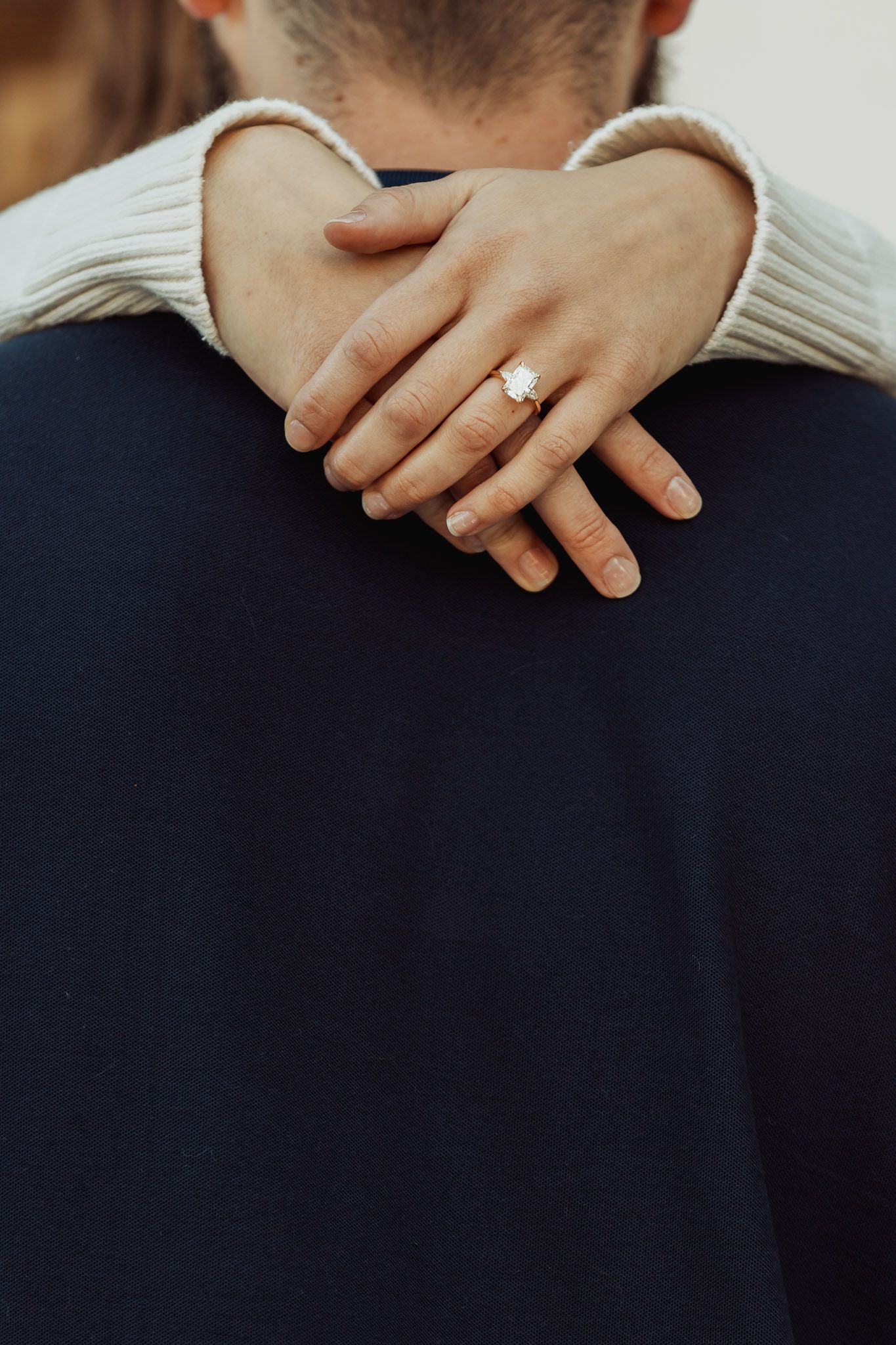 A close-up of hands wearing a white sweater and showing off an engagement ring against a dark suit jacket.