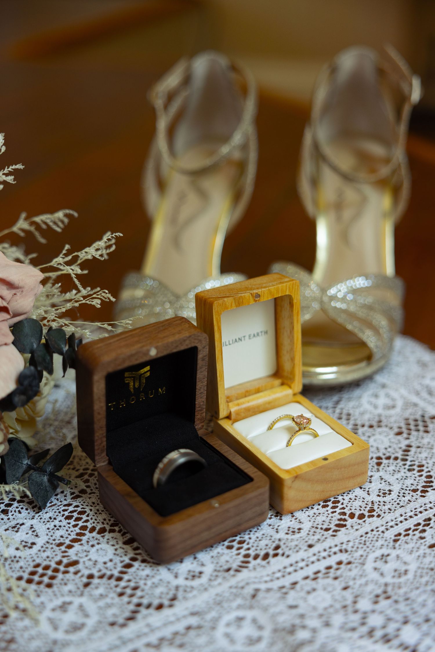 Two wooden wedding ring boxes with gold and silver bands displayed on lace beside a floral bouquet and gold heeled shoes.