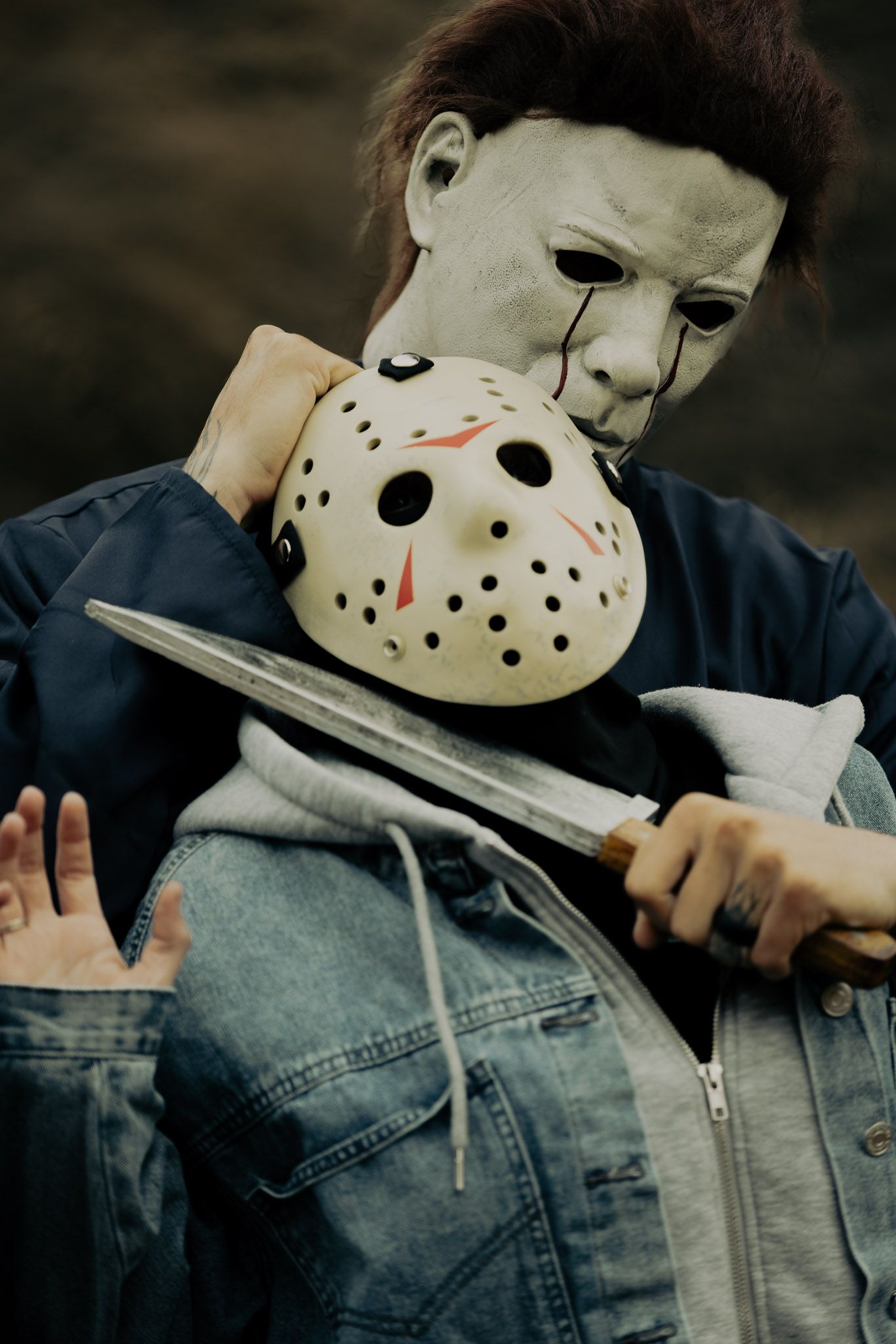 A figure wearing a hockey mask holds a machete while wearing a denim jacket in a dark setting.