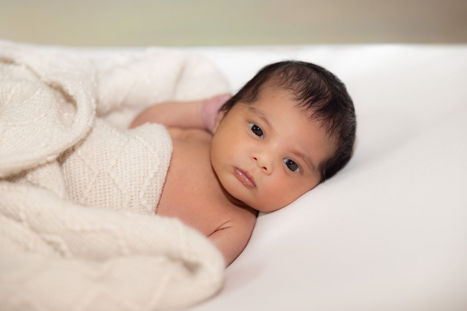 A newborn baby lies peacefully under a soft white blanket while looking up at the camera.