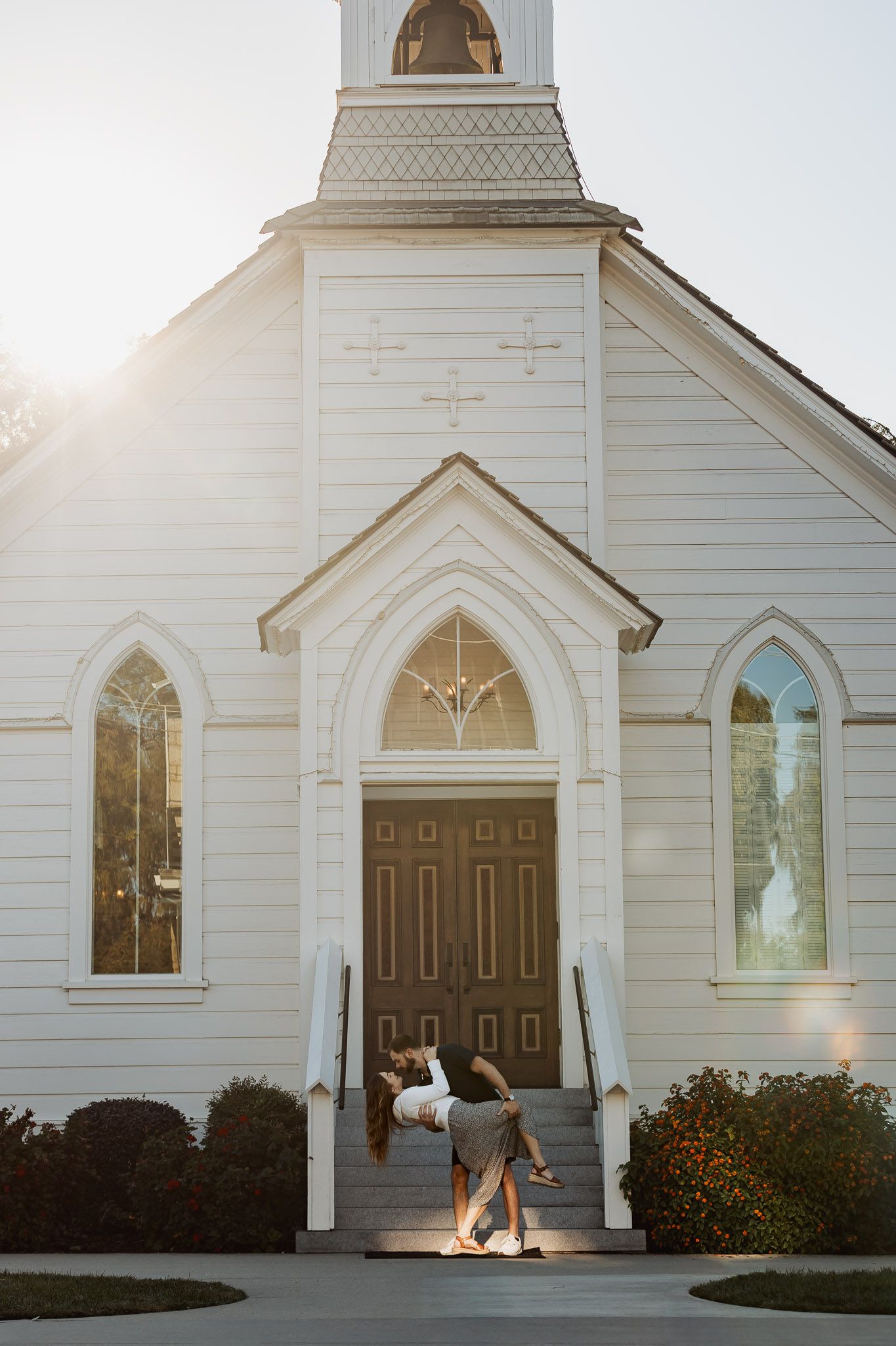A classic white church with gothic windows and a brown door features two people sitting on the front steps at sunset.