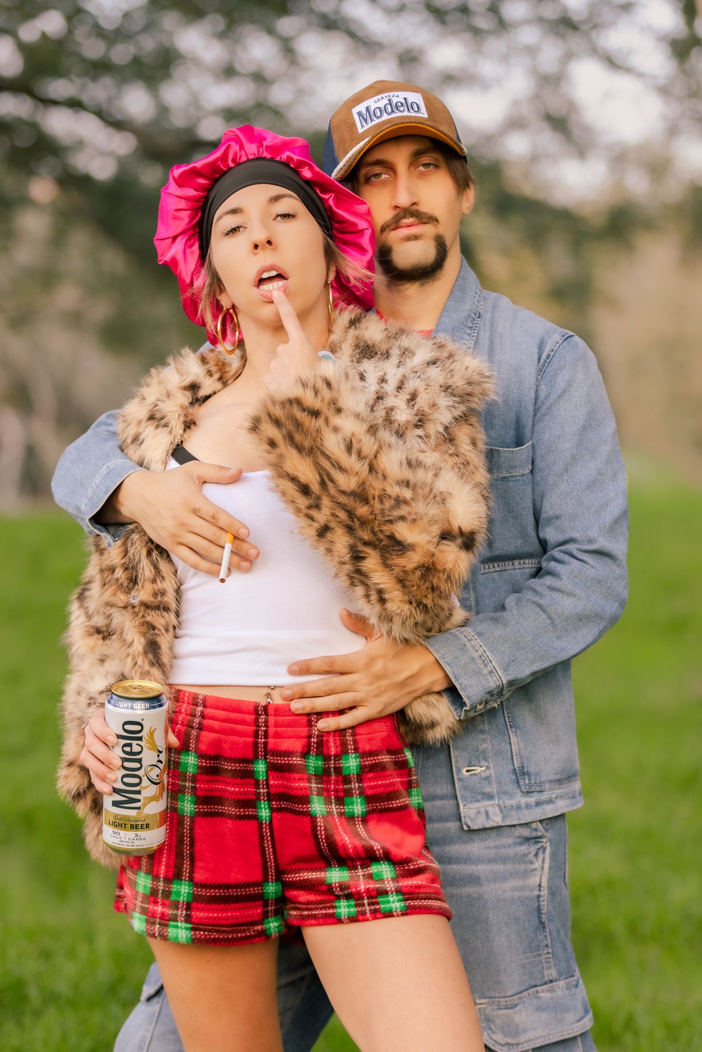 A couple embraces playfully outdoors wearing casual attire with plaid shorts and denim jacket in a grassy field.