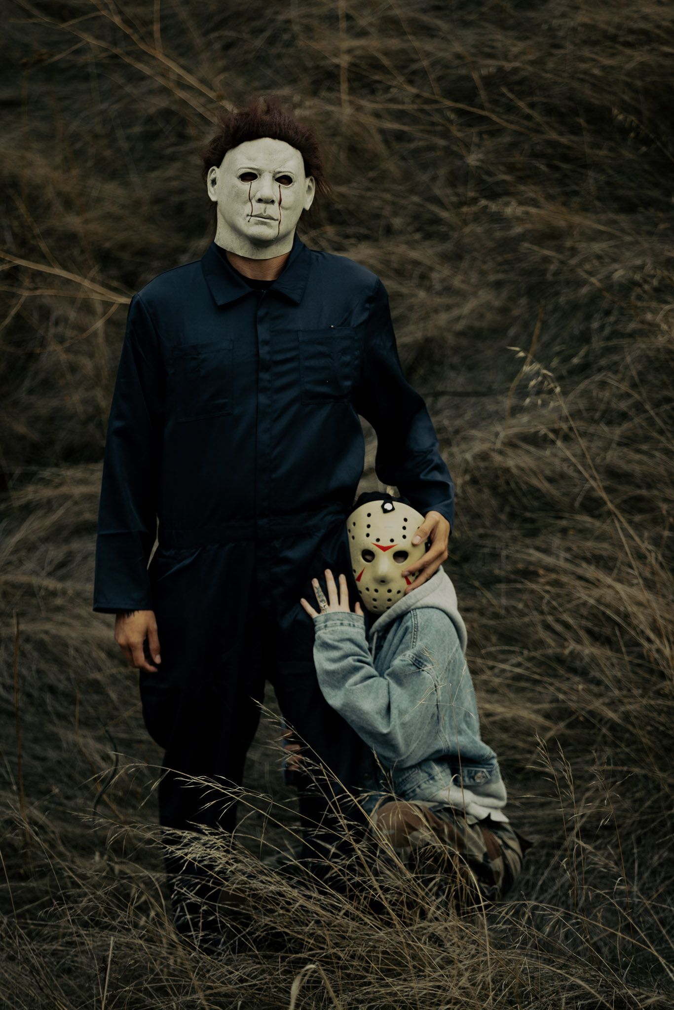 Two figures in horror movie masks stand together in a dark field wearing dark clothing.