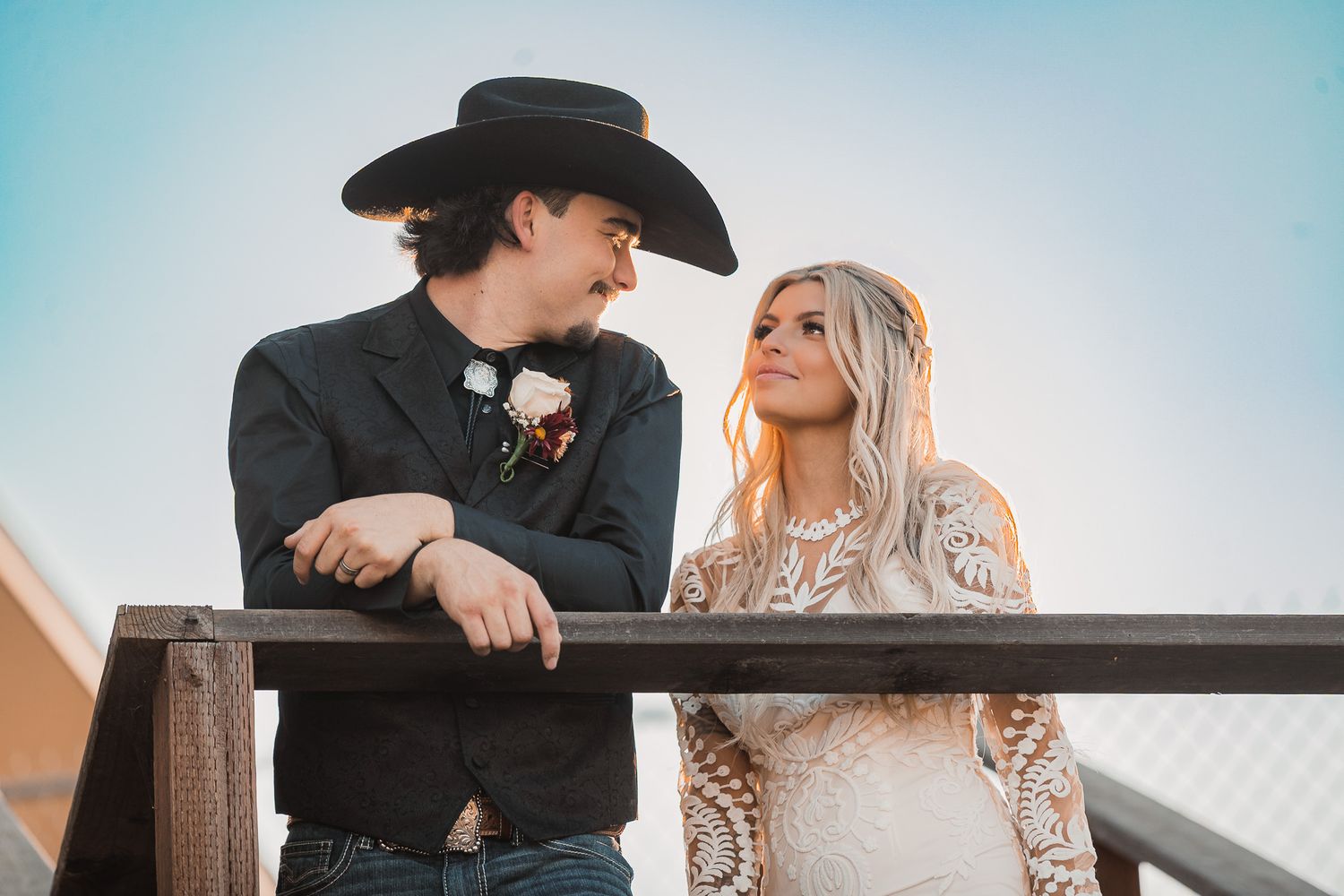 Wedding couple in western attire sharing romantic moments by wooden fence.